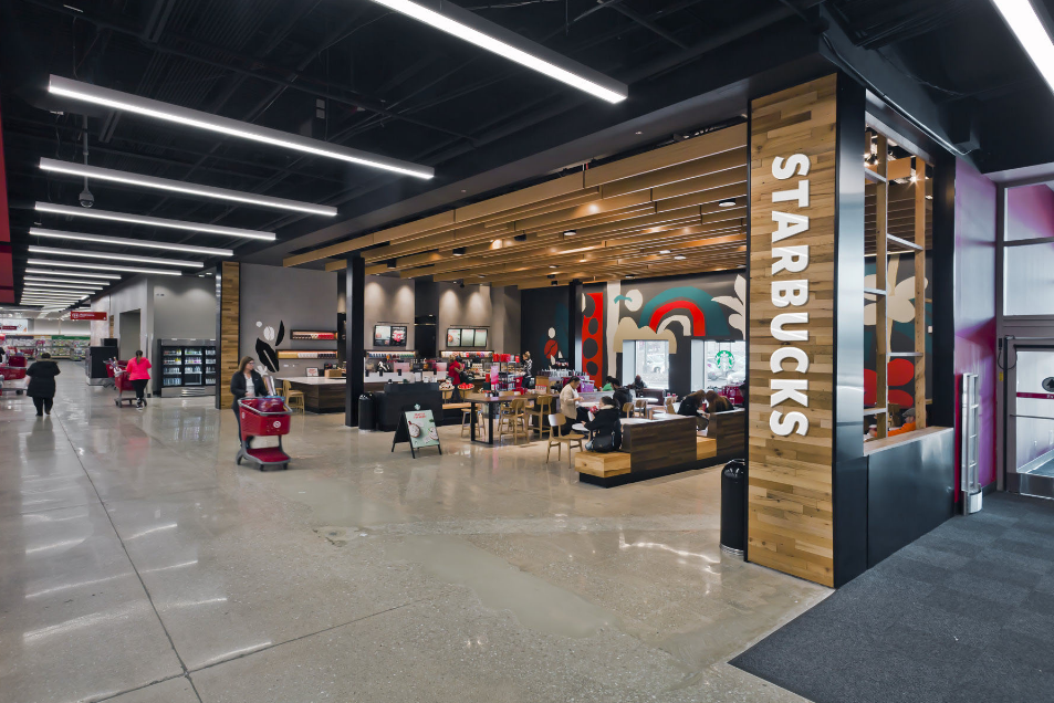 Inside a Starbucks coffee shop located in a shopping mall with customers sitting and standing, modern interior design, wooden accents, and illuminated ceiling lights.