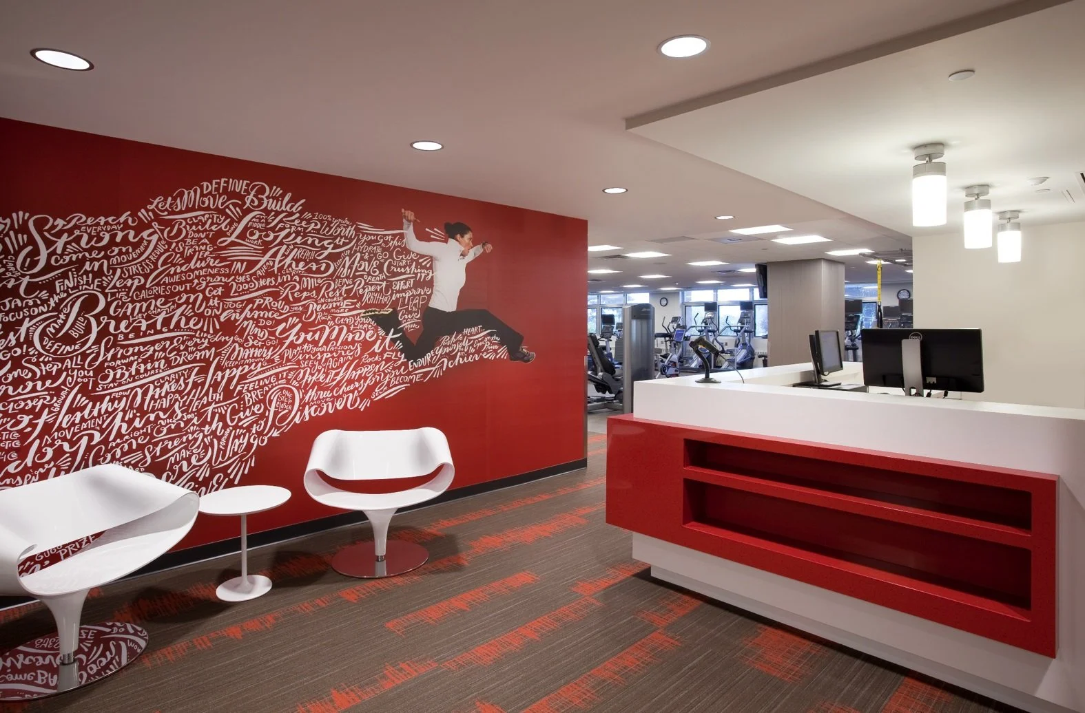 Modern gym reception area with red and white decor, abstract artwork on red wall, white chairs, and a front desk with computer monitors. Exercise equipment is visible in the background.