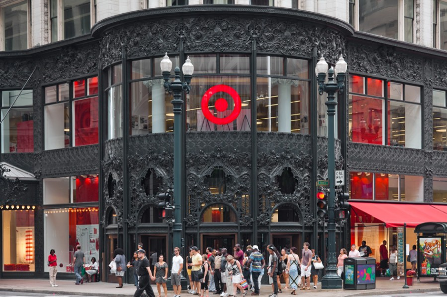 Exterior of a Target store on a busy street corner with a crowd of pedestrians in front. The store has black ornate ironwork, large glass windows, and a prominent red Target logo above the entrance.