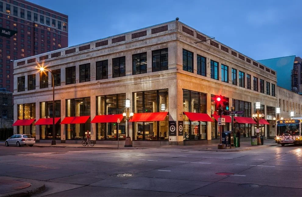 A corner building with large glass windows, red awnings, and vintage street lamps in an urban setting during dusk, with vehicles and a bus passing by.