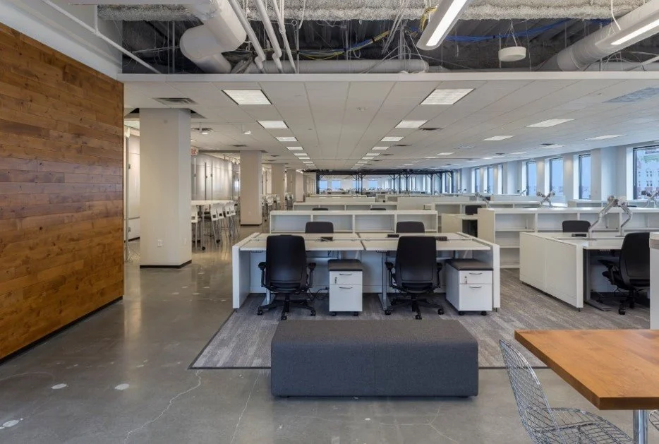 Empty modern office space with rows of workstations and chairs, large windows, and exposed ceiling pipes.