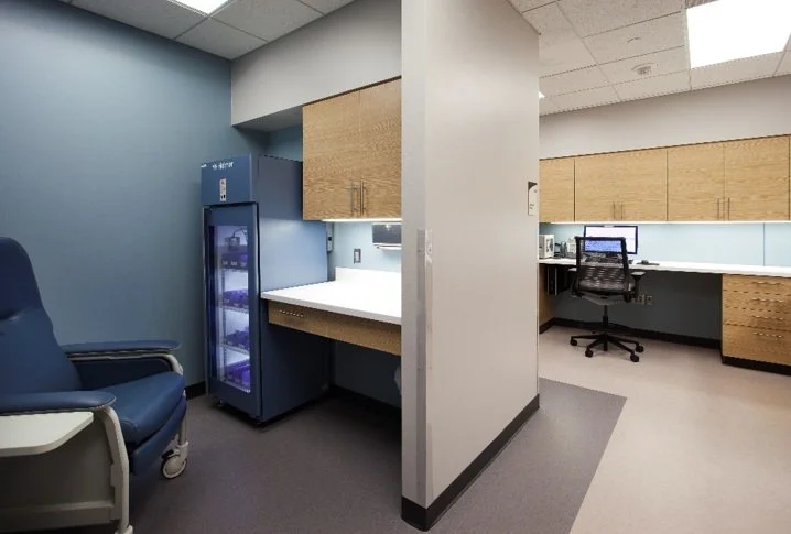 Hospital room with a blue recliner chair, a vending machine, and a workspace with a desk, office chair, and wood cabinetry.