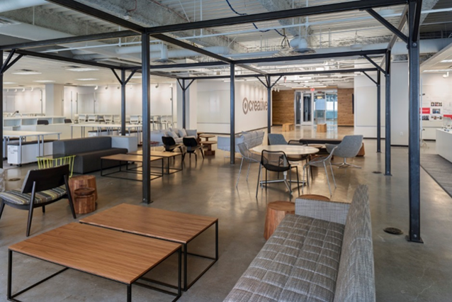 Modern office lounge with tables, chairs, sofas, and a reception area visible in the background.