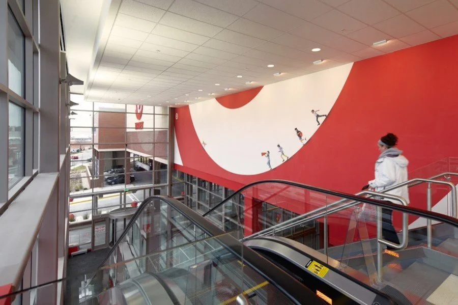 Inside view of a modern shopping mall or store with large mural wall art depicting children climbing, walking, and playing on a large, swooping red and white design. An escalator and railing are visible with a woman in a white hoodie standing nearby.