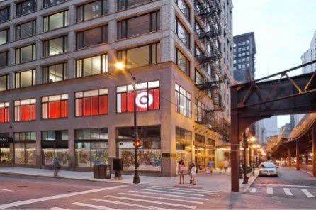 Urban street corner with a brick building featuring large windows, streetlights, pedestrians, and a car on the road during dusk in a city.