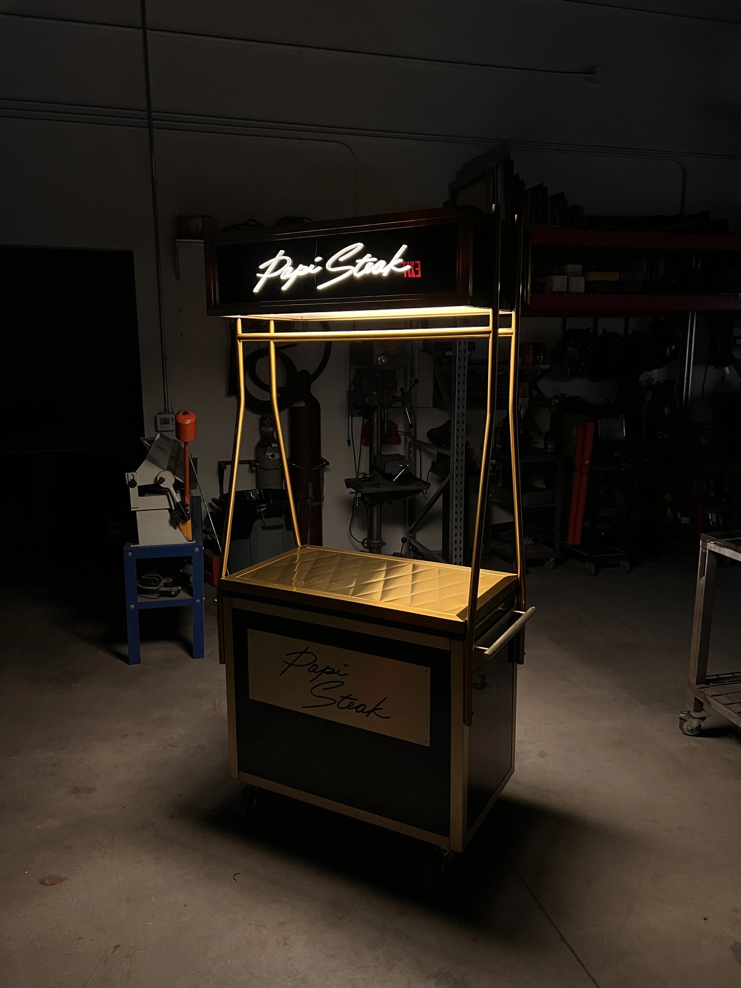 A black and gold Papí Steak food cart with illuminated signage in a dimly lit indoor setting.
