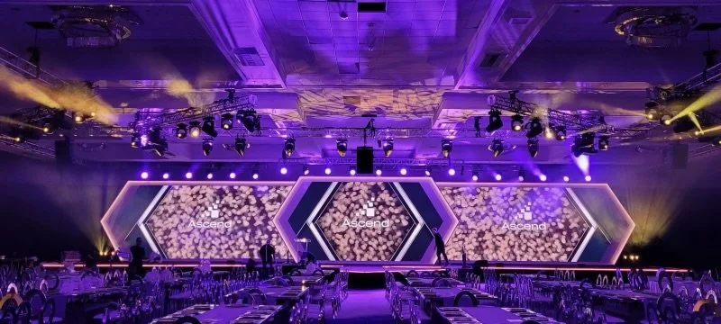 Large event hall with a stage featuring multiple hexagonal display screens showing the word 'Ascend' and decorative background, illuminated with purple and yellow lighting, with tables and chairs arranged for attendees.