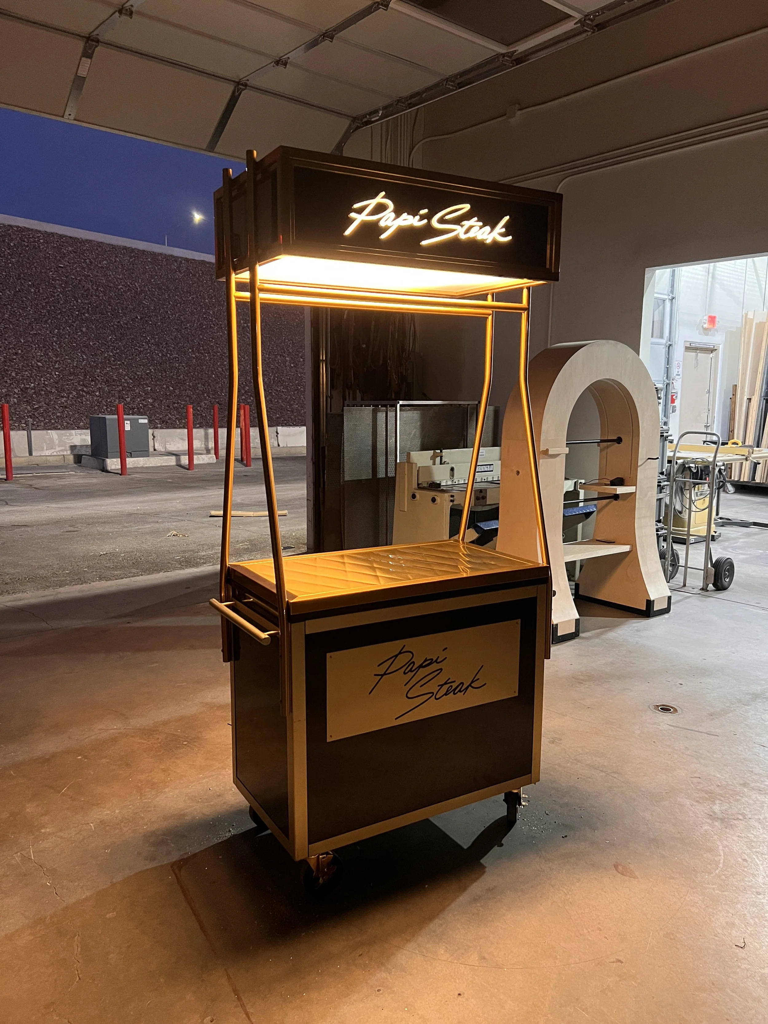 A small rolling cart with a black and yellow sign reading 'Papi Steak' in cursive, illuminated by warm lighting, positioned inside a garage or storage area.