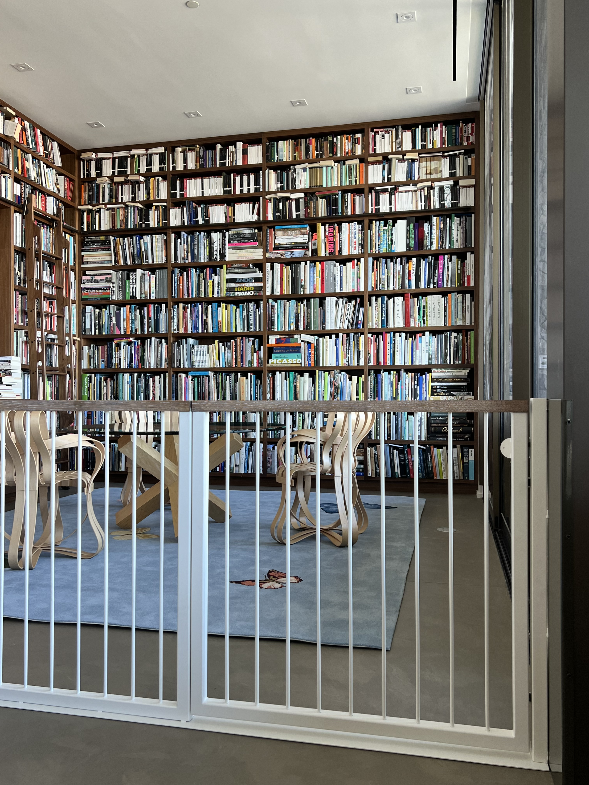 A large wooden bookshelf filled with books, a rug with a red fox illustration, wooden chairs with curved backs, and a small wooden table, in a well-lit room with a glass wall or window.