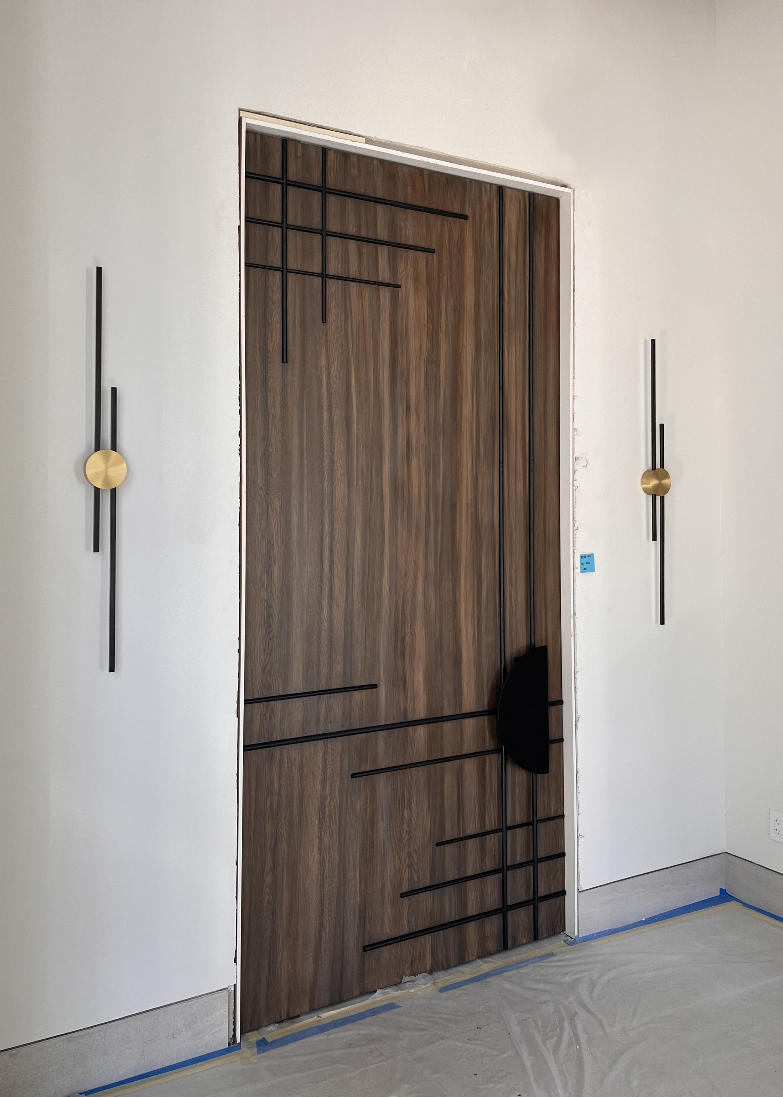 Interior door with geometric black lines design, flanked by two modern wall sconces with black rods and gold discs, on a white wall.