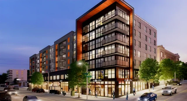 Modern multi-story apartment building with glass balconies, situated on a city street with cars and pedestrians, during dusk.