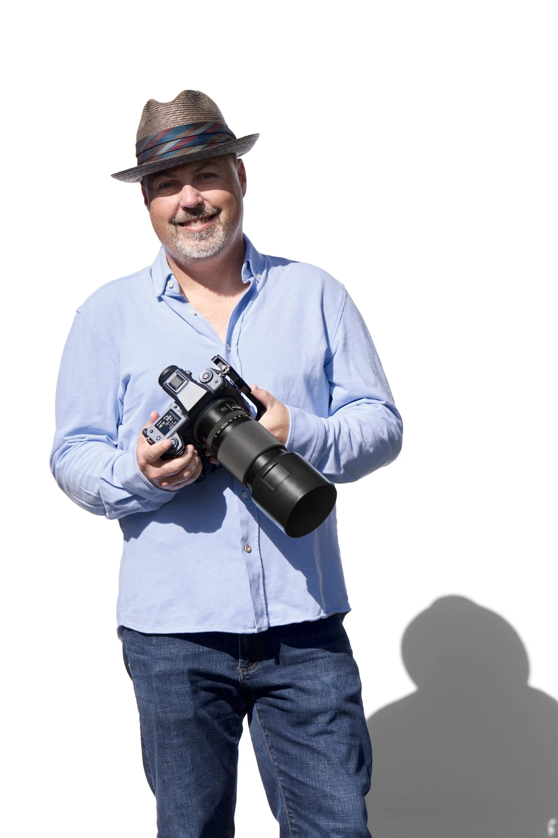 A man in a light blue button-up shirt and jeans holds a camera with a large telephoto lens. He wears a fedora hat with a colorful band and smiles at the camera, standing against a plain white background.