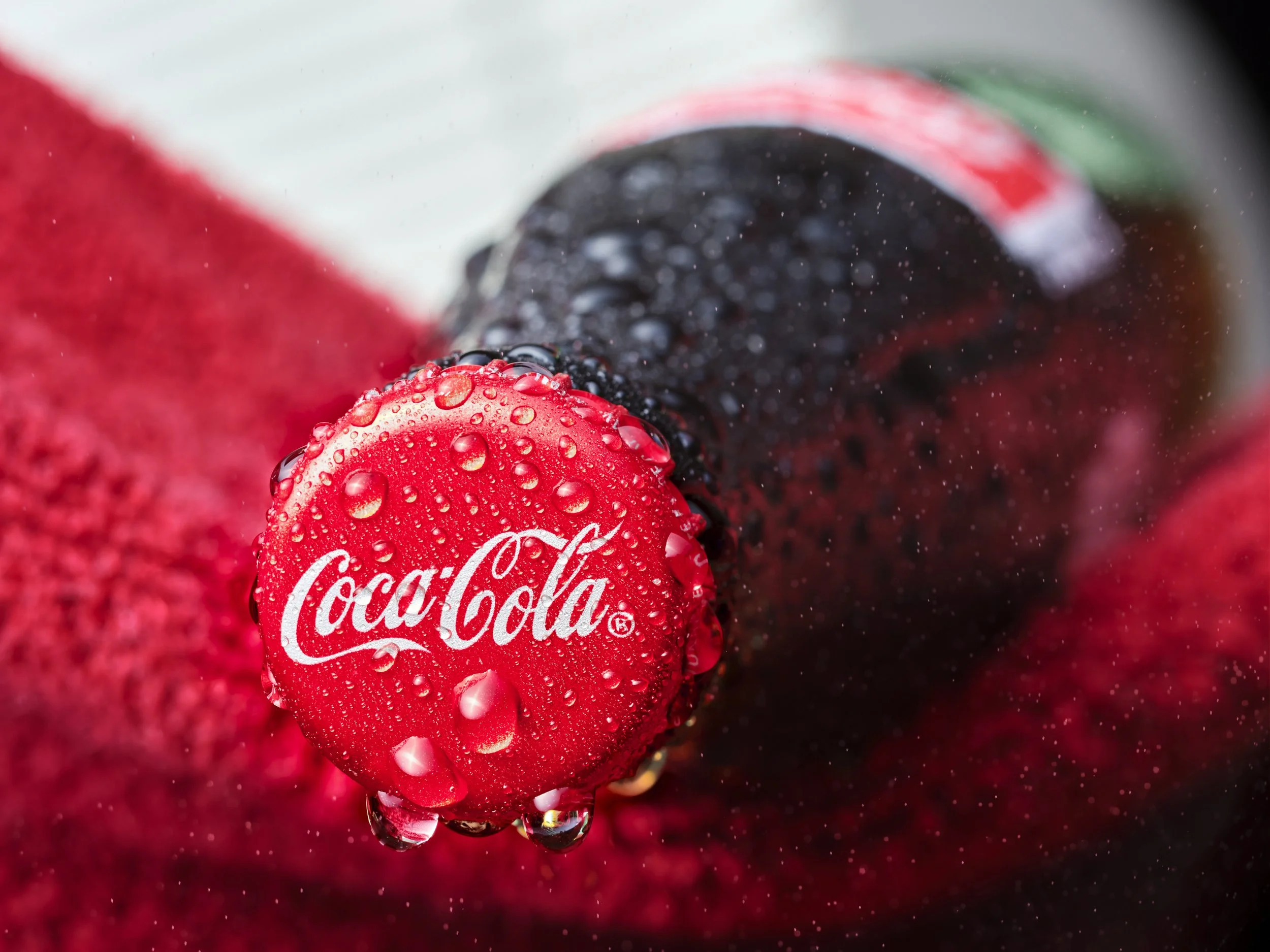Close-up of a red Coca-Cola bottle cap with water droplets on it, placed among black and red berries, with a soft focus background.