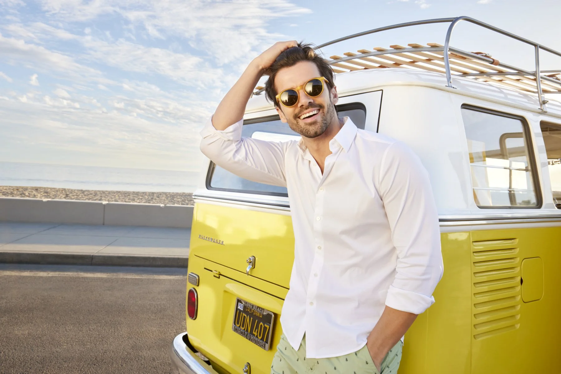 A man wearing sunglasses and a white shirt smiling and posing with one hand on his head, standing next to a yellow vintage van parked by the beach with ocean and sky in the background.