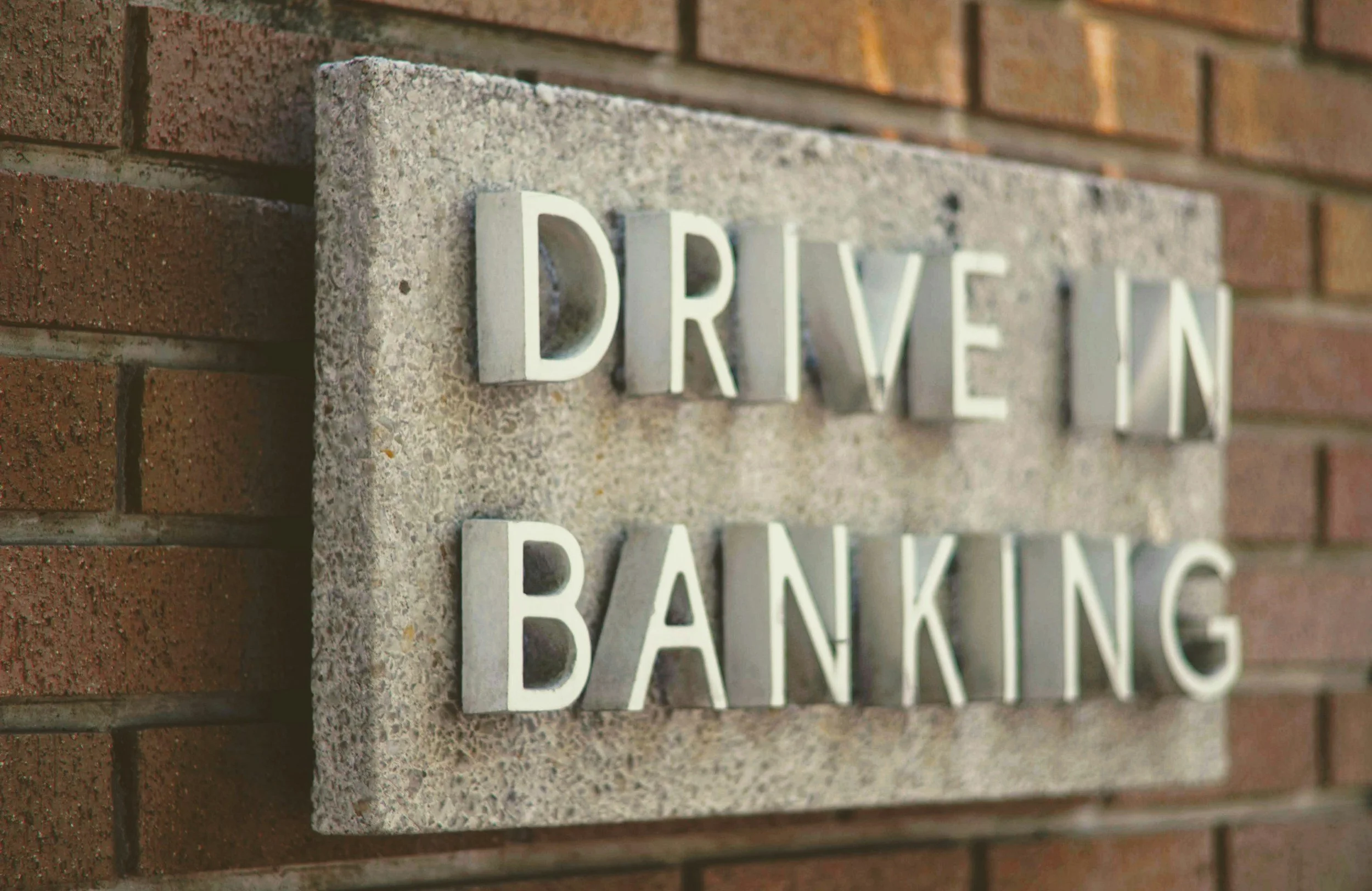 Sign on brick wall reading "Drive In Banking" with metal letters on a concrete background.
