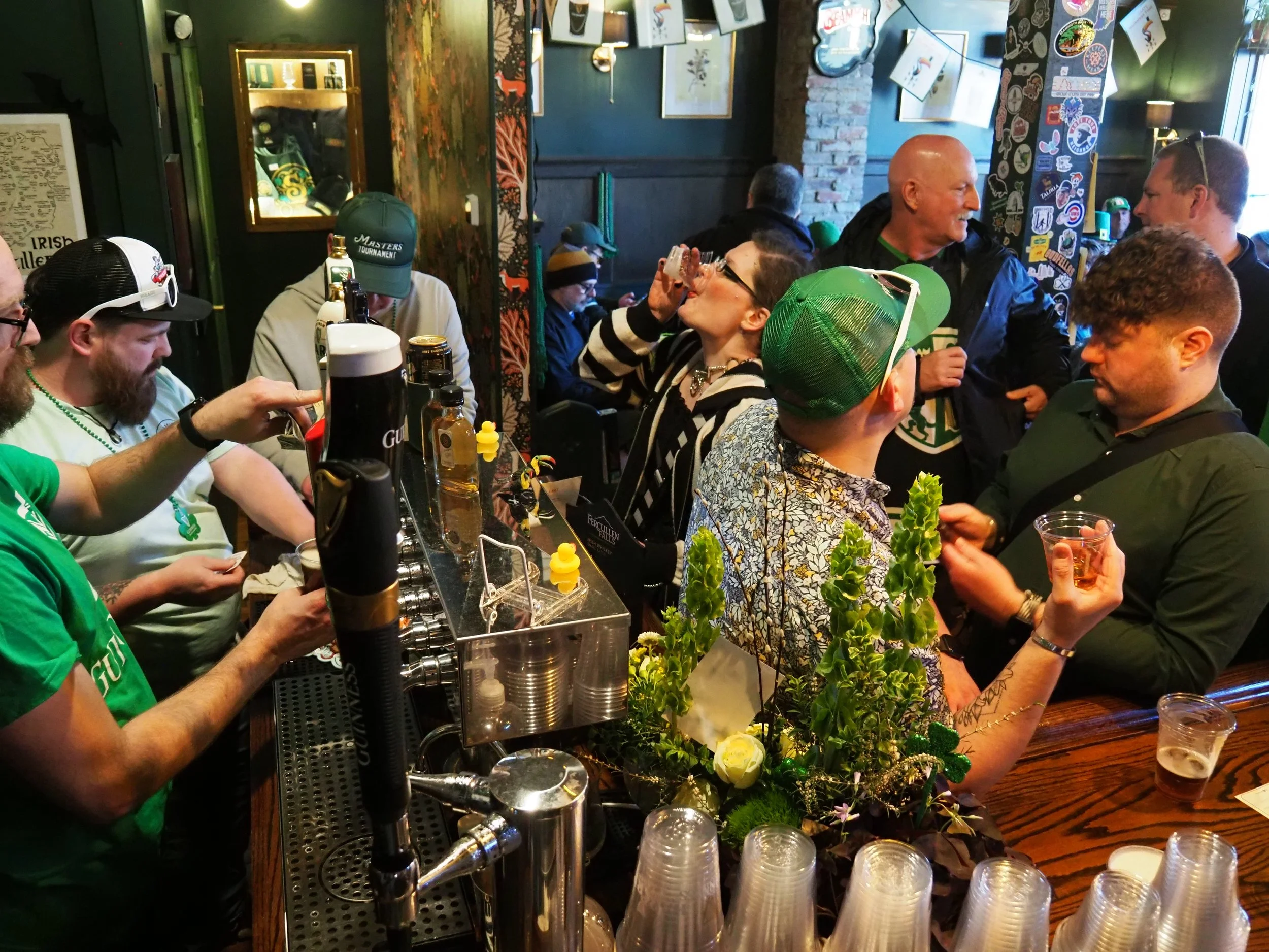 People gathered at a bar, drinking and socializing, with a woman in striped shirt and glasses drinking from a shot glass. The bar is decorated with plants and various stickers on a pillar.