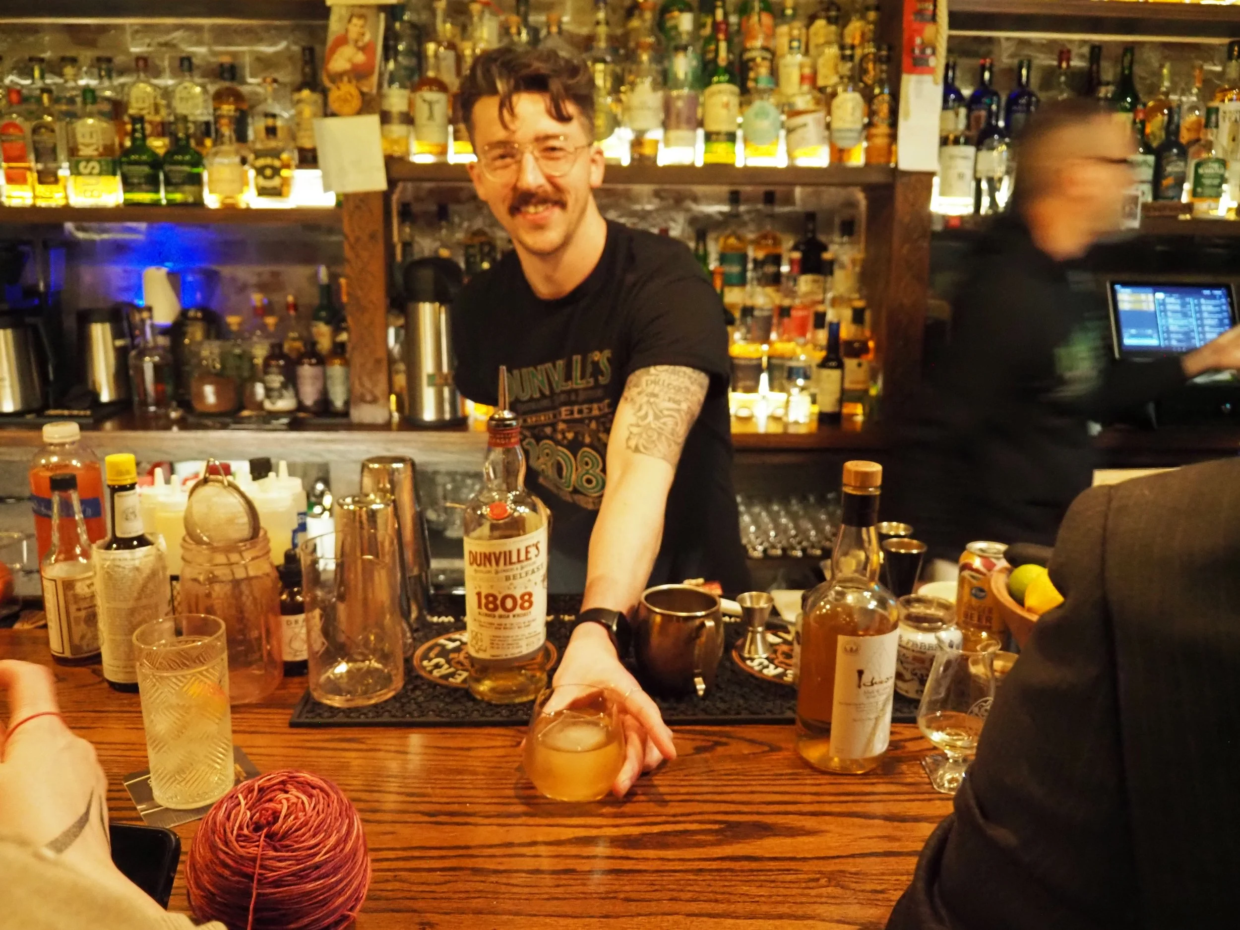 A bartender with tattoos and glasses serving a drink at a bar with various bottles of liquor in the background, and a person in a suit partially visible on the right.