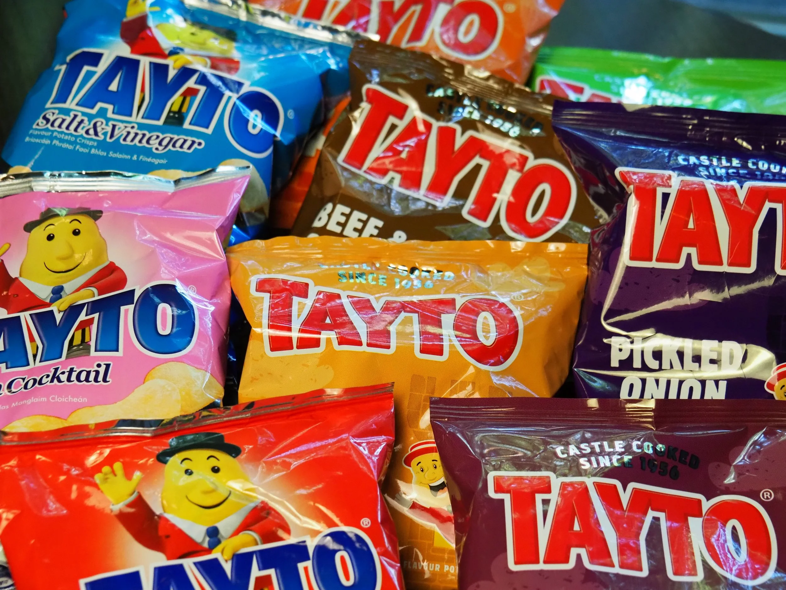 A colorful assortment of TAYTO snack bags including salted vinegar chips, beef crisps, cocktail crisps, and pickled onion flavors.