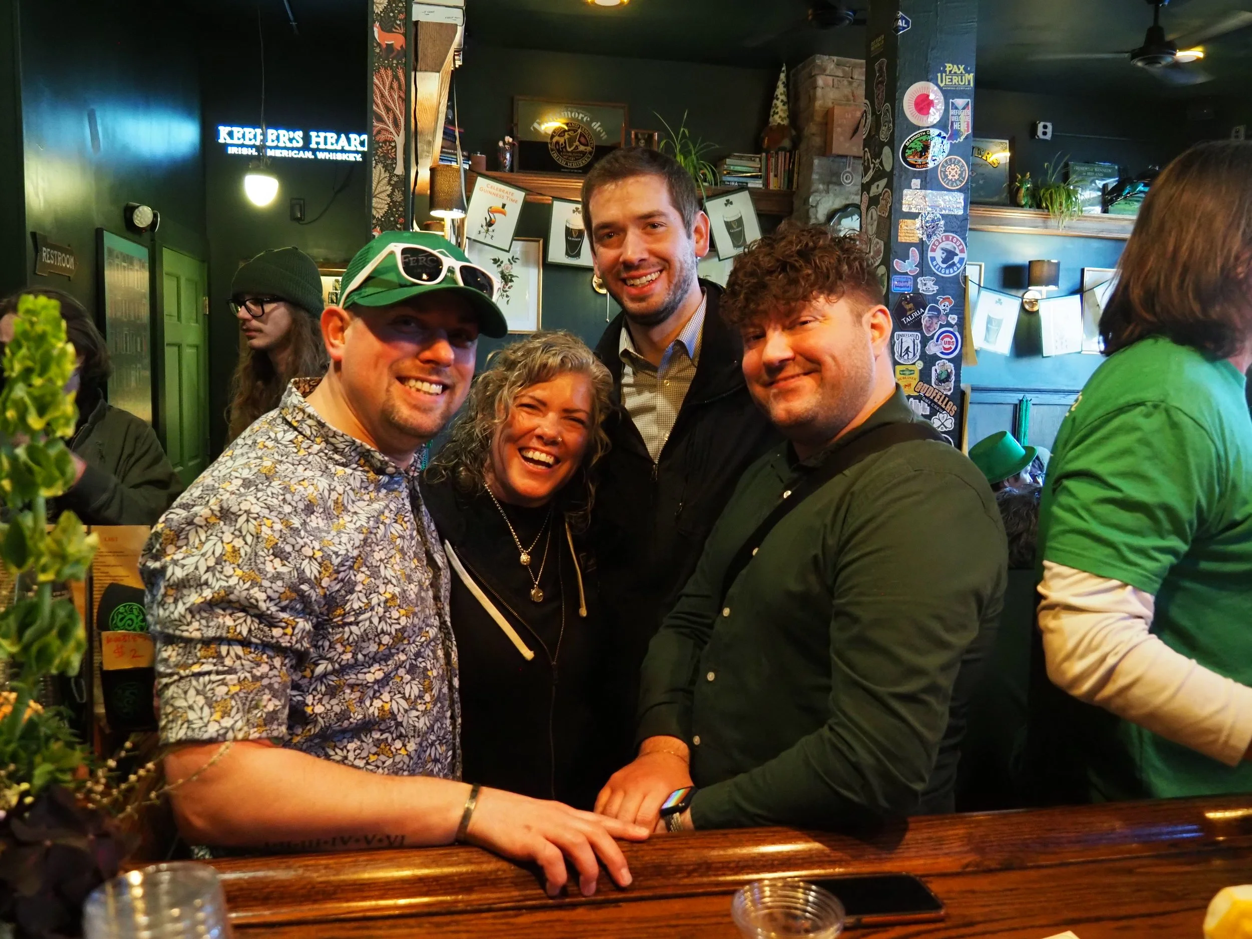 A group of five people smiling and posing together in a bar or pub. Four men and one woman, with some wearing casual and colorful clothing, are standing close to each other behind a wooden bar counter.