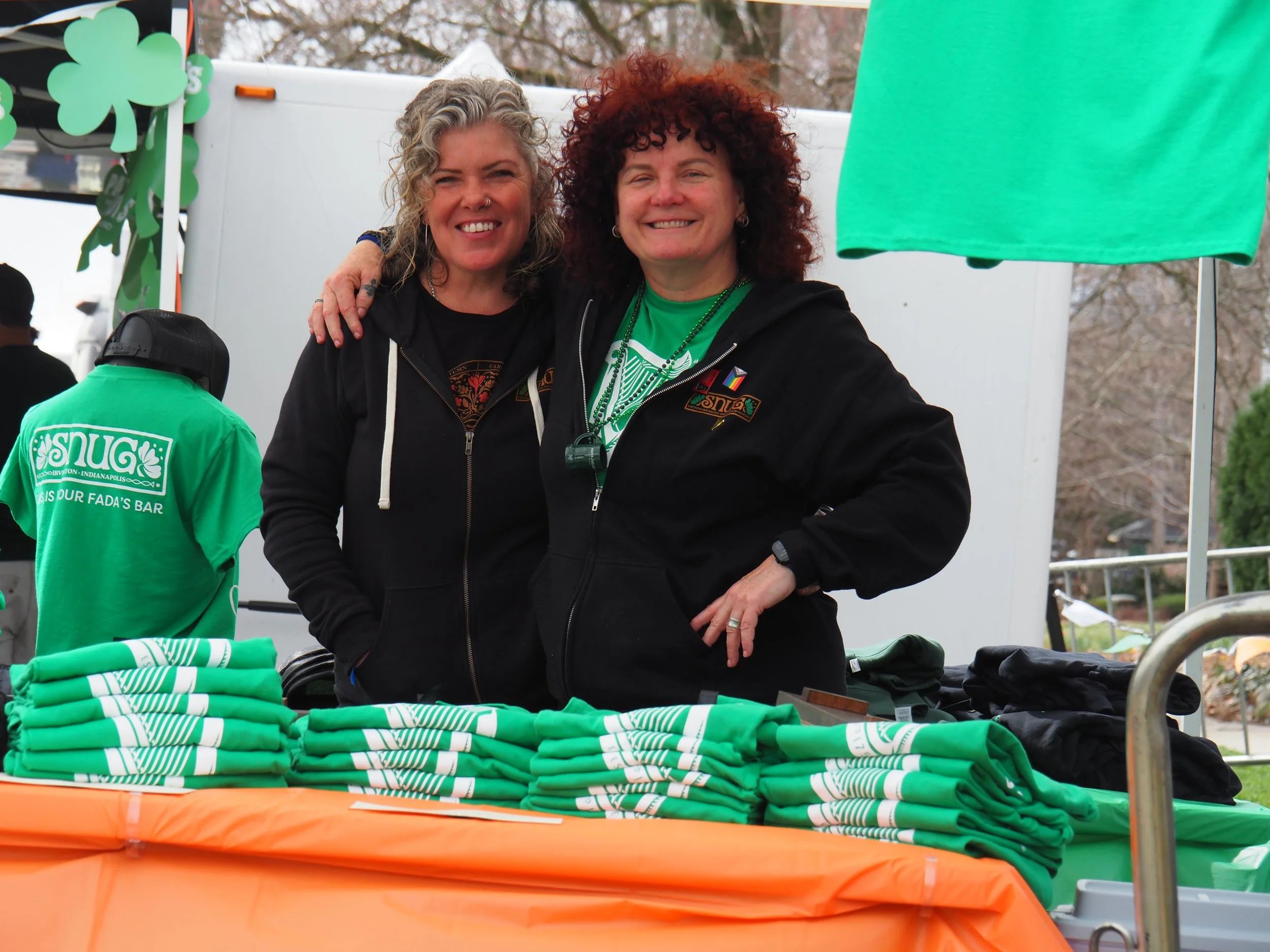 Two women smiling behind a table with green t-shirts folded on top at Snug St. Patrick's Day 2025
