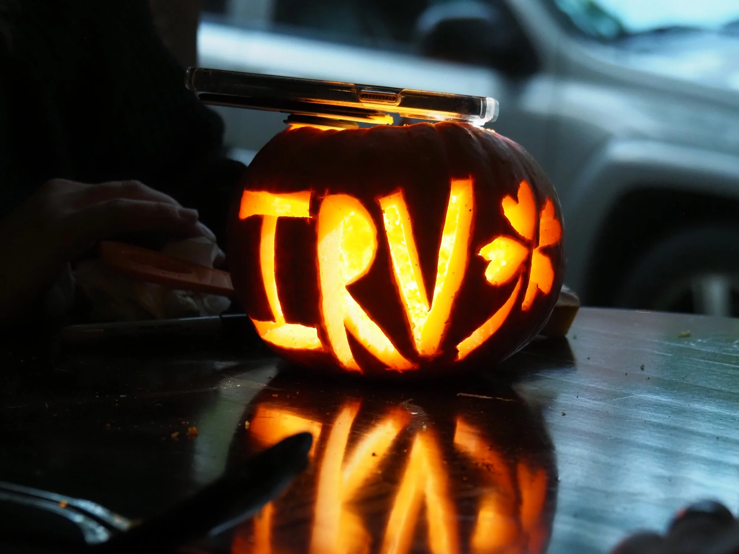 A carved pumpkin with the word "TRIV" and a heart and butterfly design, illuminated from within, placed on a reflective surface in a dark room with a mobile phone resting on top.