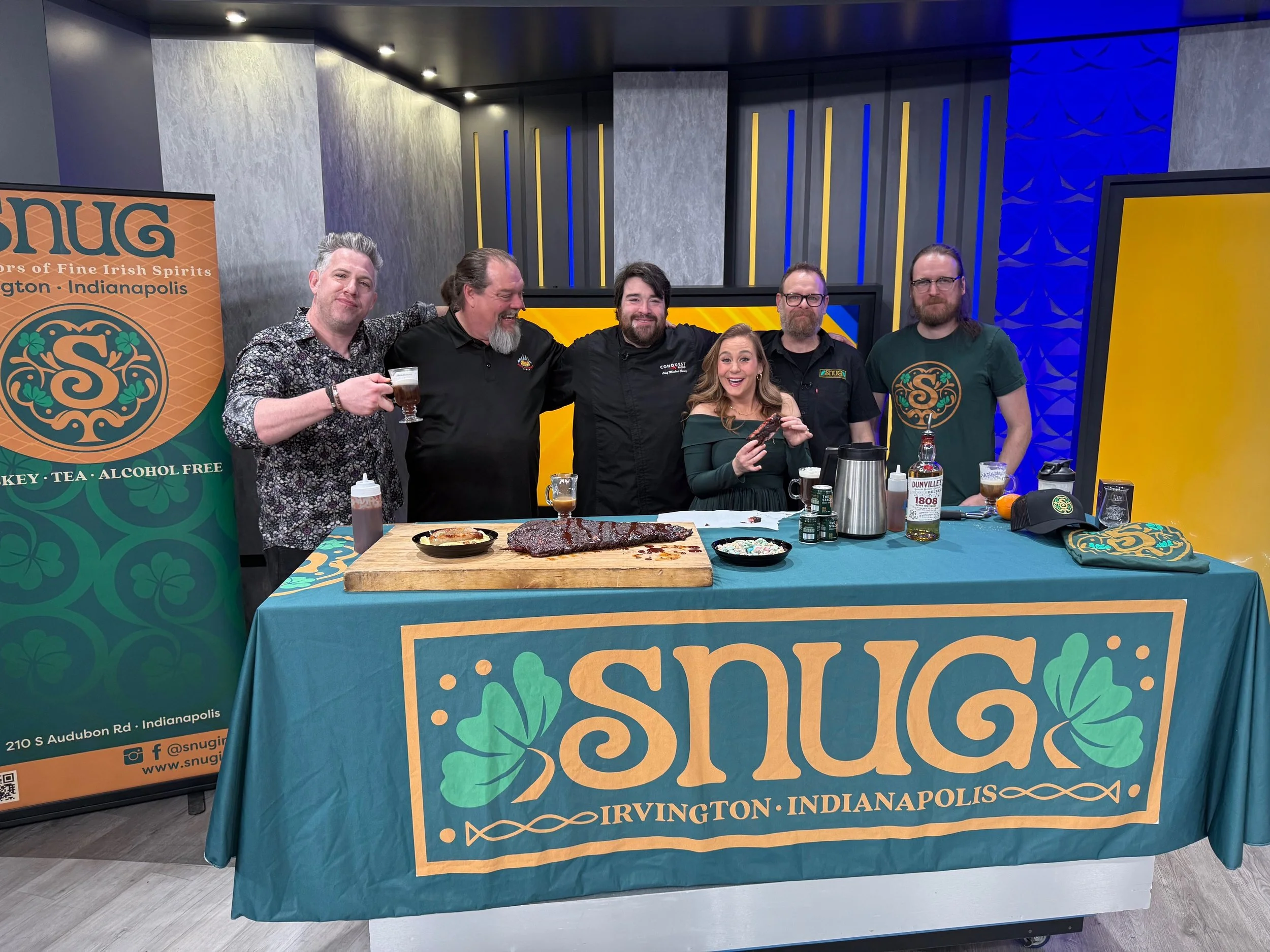 Group of six people standing behind a table with food and drinks, celebrating Snug Irish pub at a TV station in Indianapolis.
