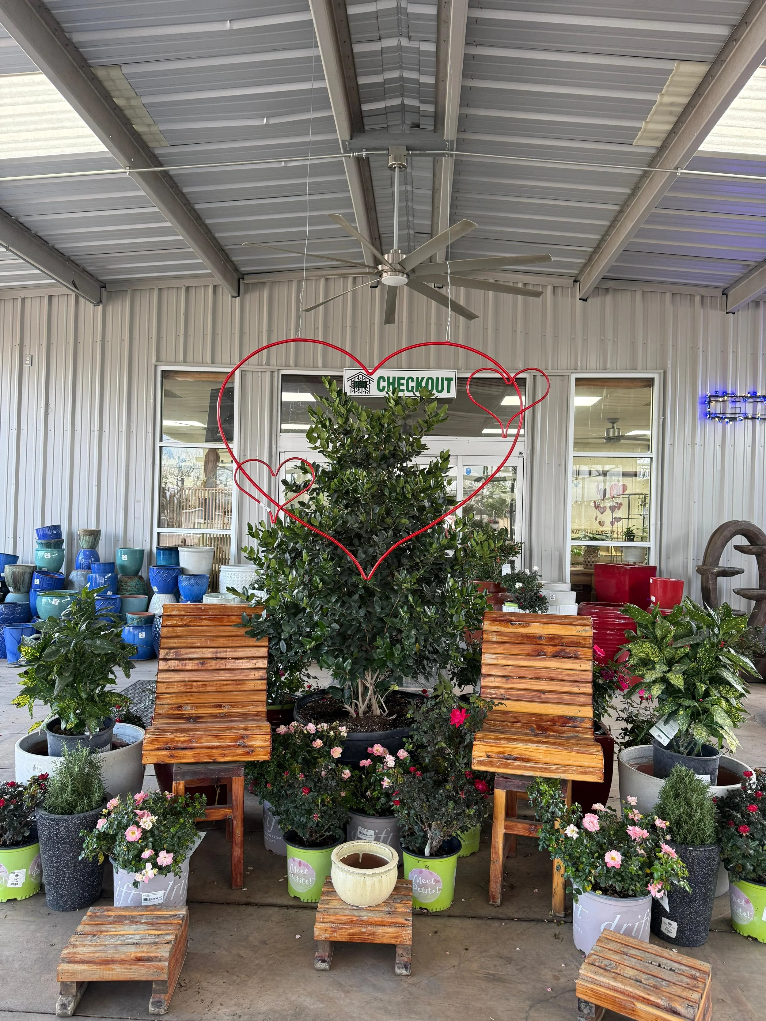 Decorative display with potted plants and wooden furniture inside a store, featuring a large bush with a red neon heart-shaped outline and smaller hearts, an 'EXIT' sign, and a glass door leading outside.