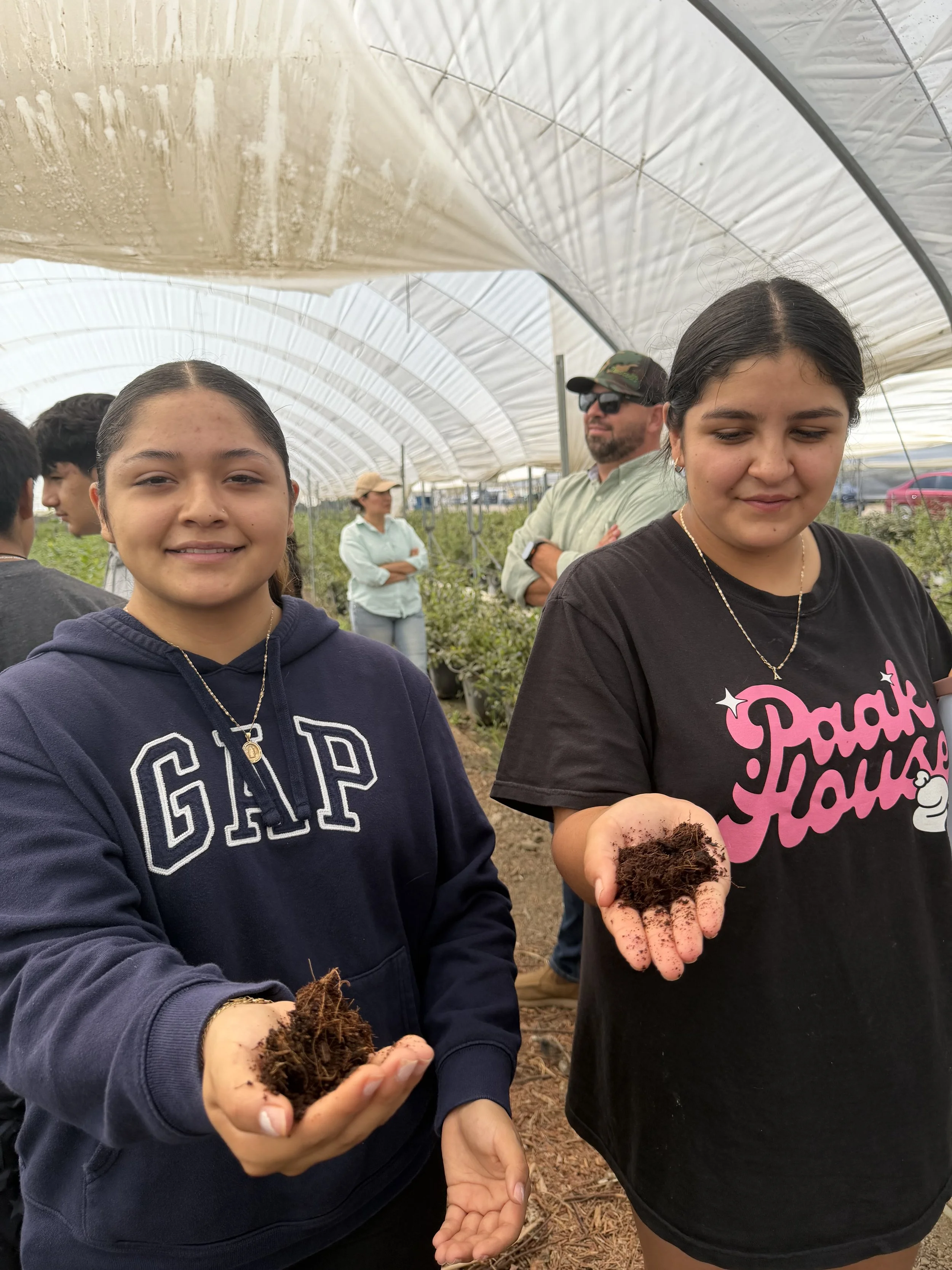 Two young girls holding soil-covered bulbs in a greenhouse, with other people in the background.