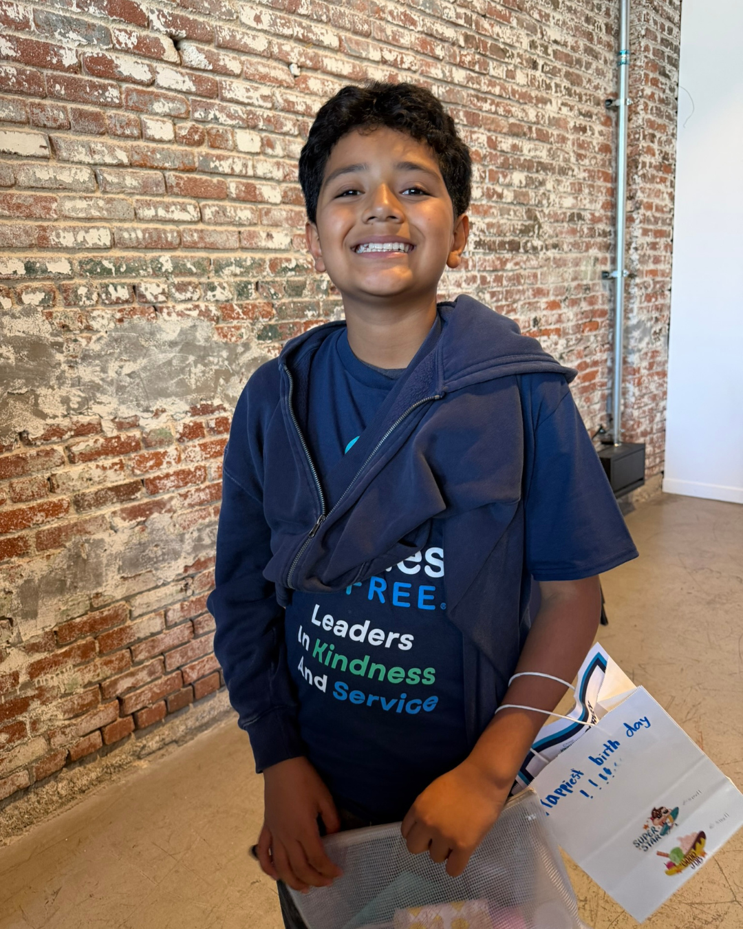 Young boy smiling, holding a gift bag, wearing a navy blue jacket and a T-shirt with words about leadership and kindness, standing in front of a brick wall, celebrating a birthday.