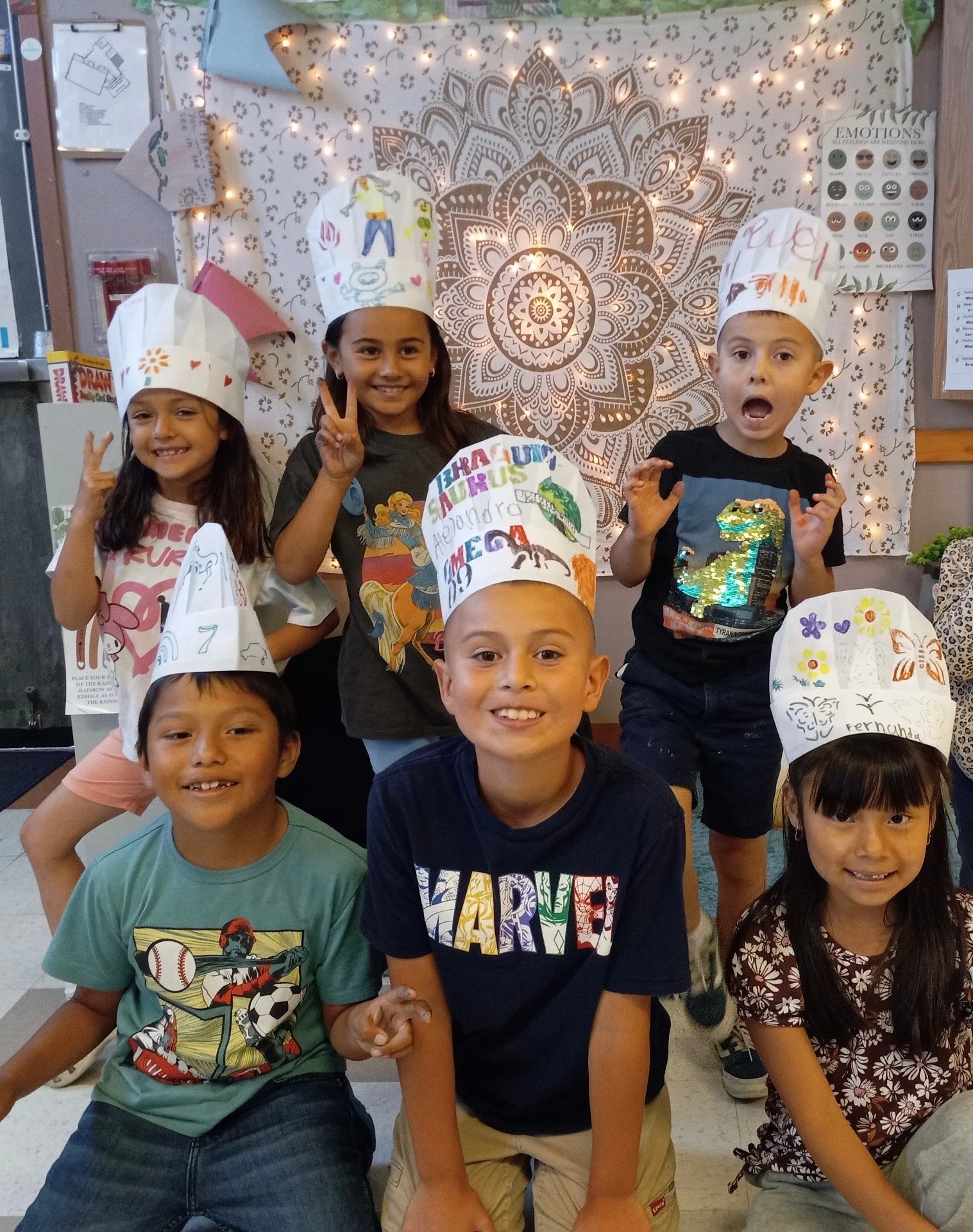 Group of children wearing chef hats, smiling, and posing for a photo in front of a decorative wall with lights, at a celebration or event.