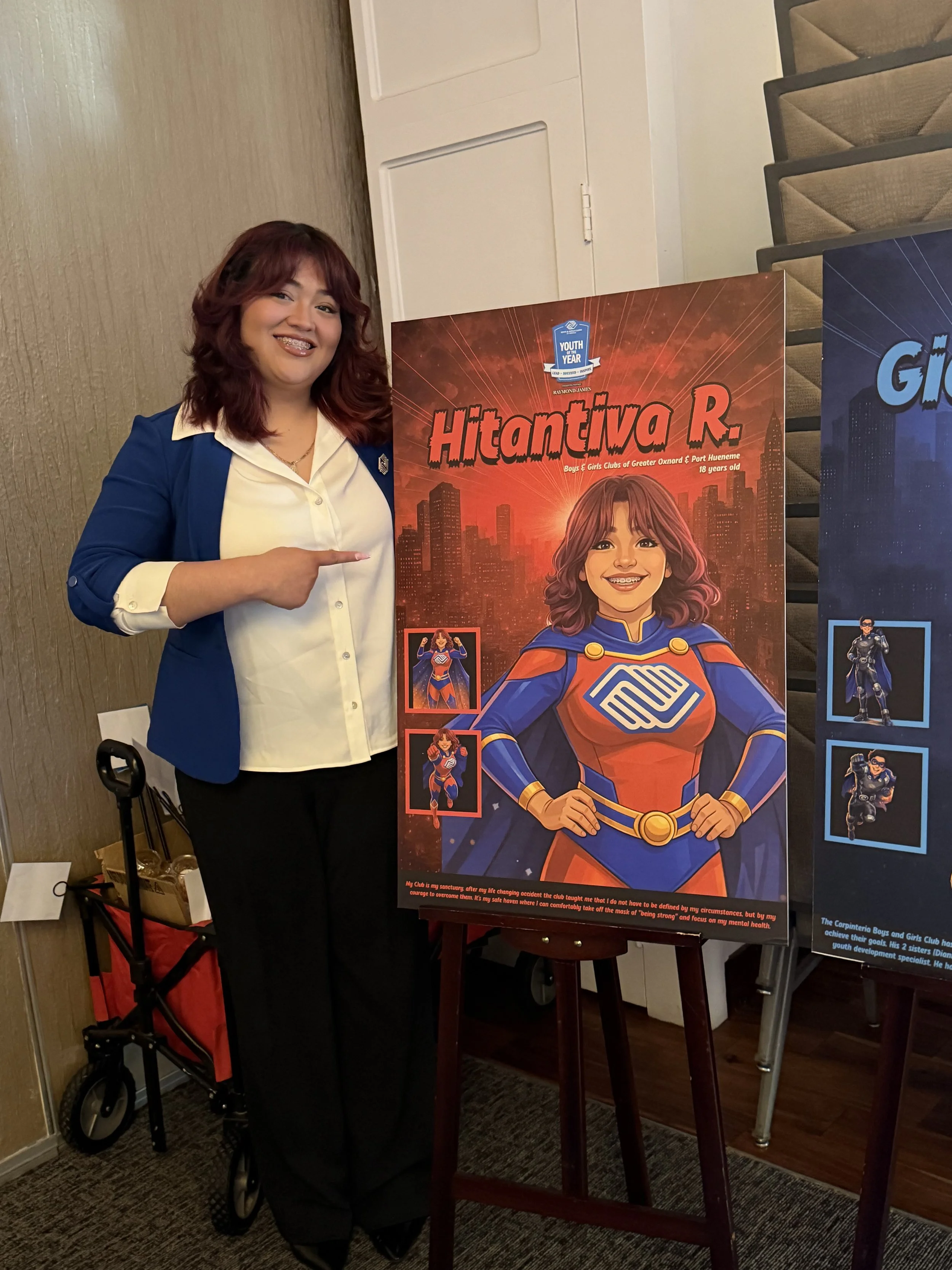 A woman with red hair and glasses standing next to a colorful poster of a young girl dressed as a superhero, pointing at the poster with her right hand.