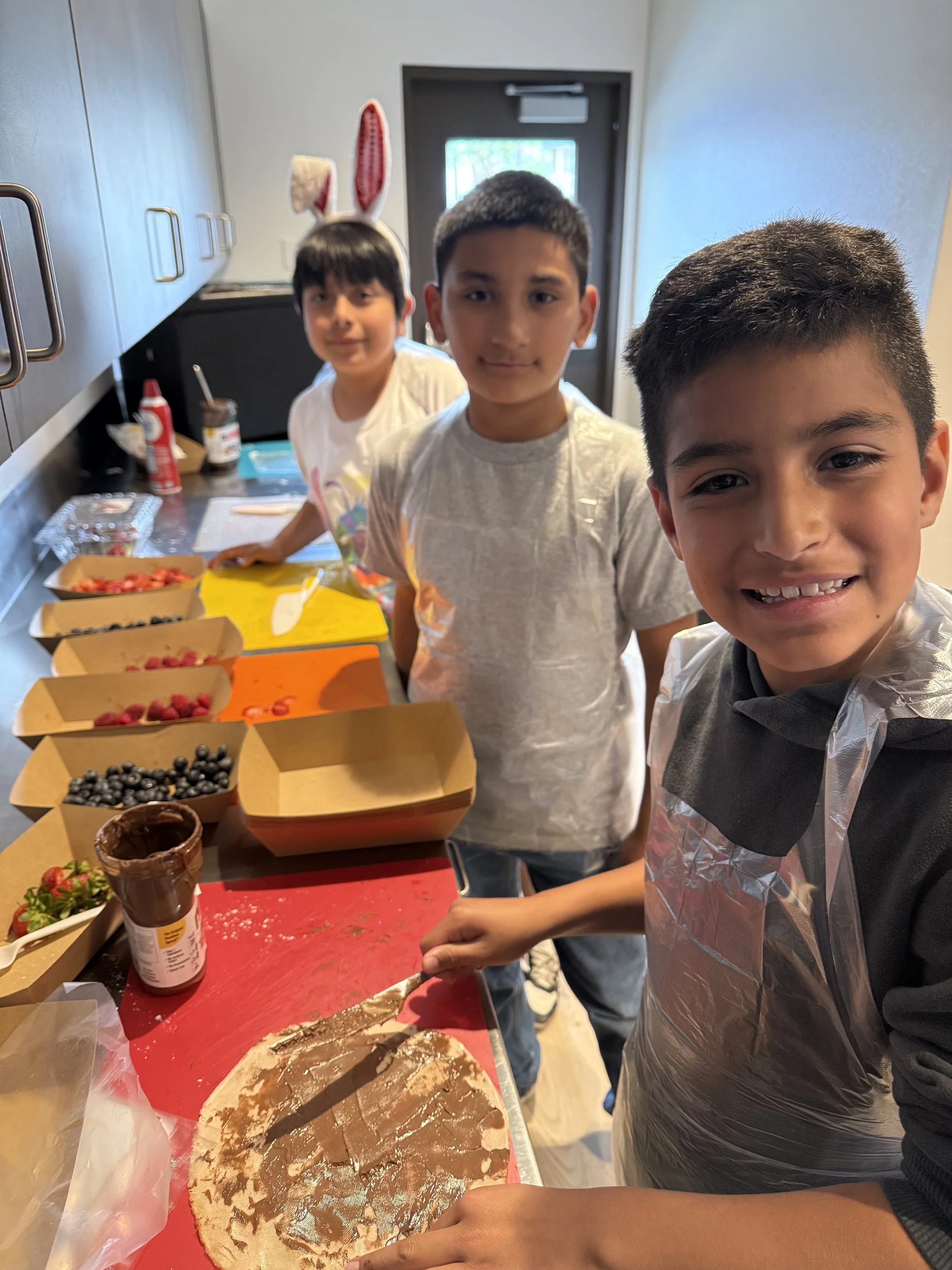 Three boys in a kitchen, one with bunny ears, standing by a table with various food items and a partially decorated cake.
