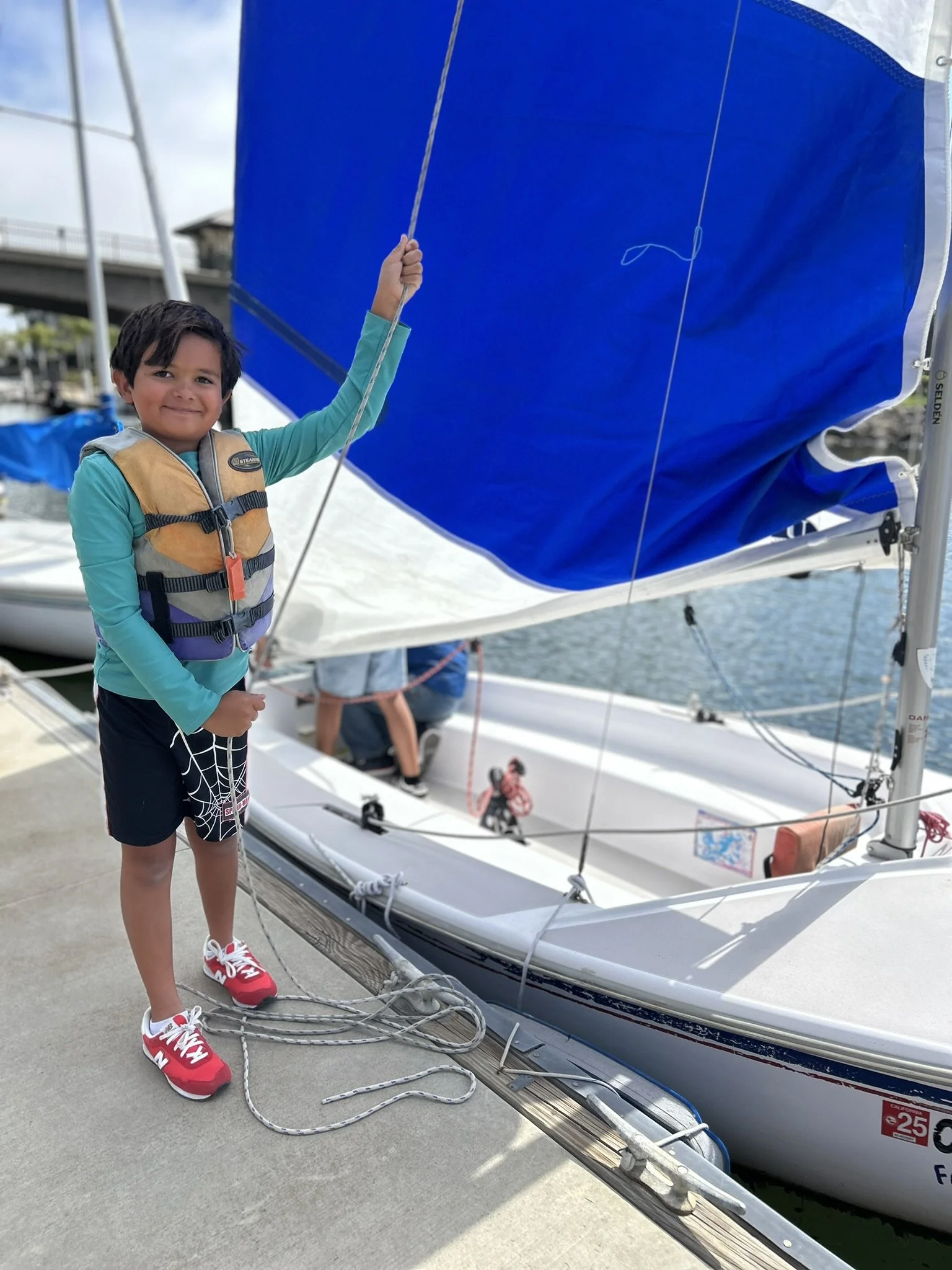A young boy in a life jacket and colorful sneakers standing on a dock next to a small sailboat, holding a rope with water and a blue sky in the background.