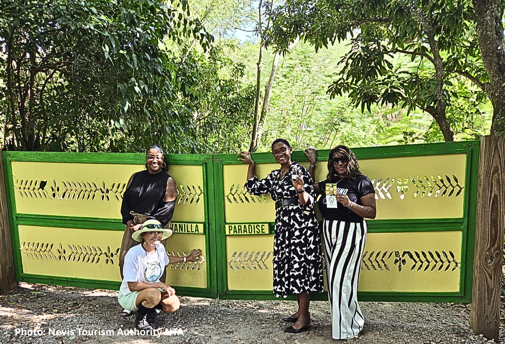 Four women standing in front of a green decorative fence with leafy trees in the background, smiling and posing for a photo at a theme park or zoo entrance.