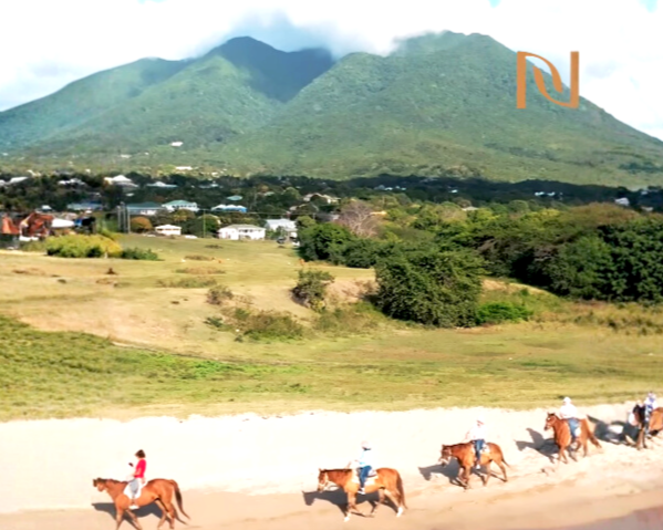 Group of people riding horses on a dirt trail with green mountains in the background and houses below.