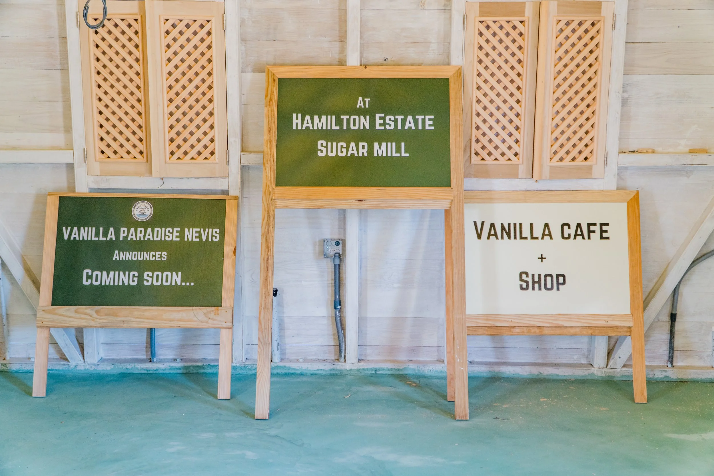 Three signs on wooden easels inside a light-colored wooden structure. The left sign states 'VANILLA PARADISE NEVIS ANNOUNCES COMING SOON...'. The middle sign states 'AT HAMILTON ESTATE SUGAR MILL'. The right sign states 'VANILLA CAFE + SHOP'.