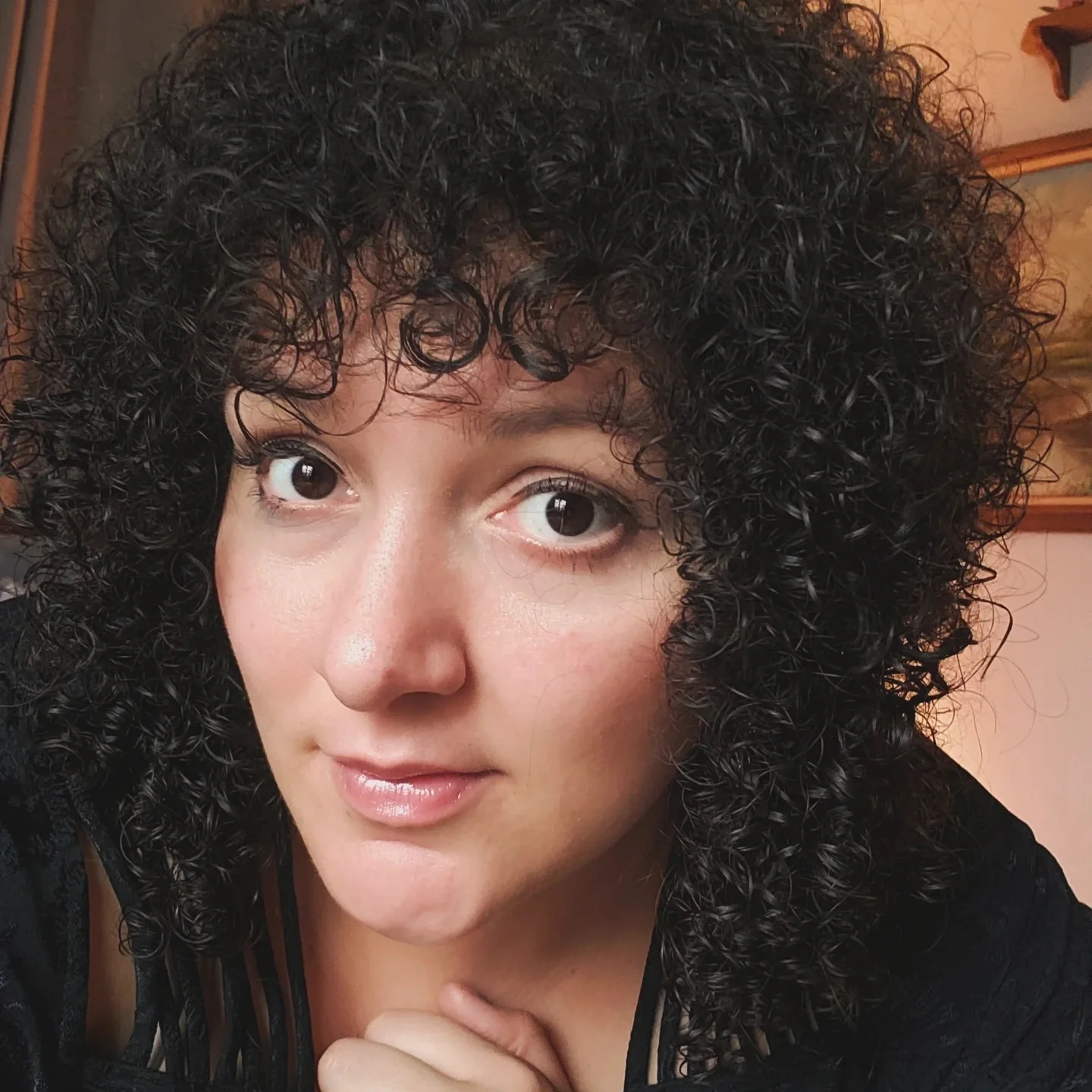 A woman with short, curly black hair. She has fair skin, and striking dark eyes, and is wearing light makeup with natural lipstick. She appears to be indoors with decor in the background.