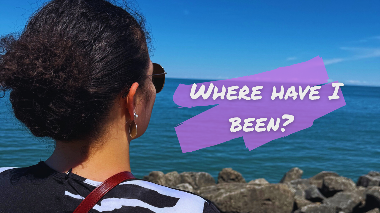 A woman with curly dark hair, wearing sunglasses, earrings, and a zebra print shirt, looks out at the ocean with rocks in the foreground. Text overlay says, "Where have I been?" in purple speech bubble.