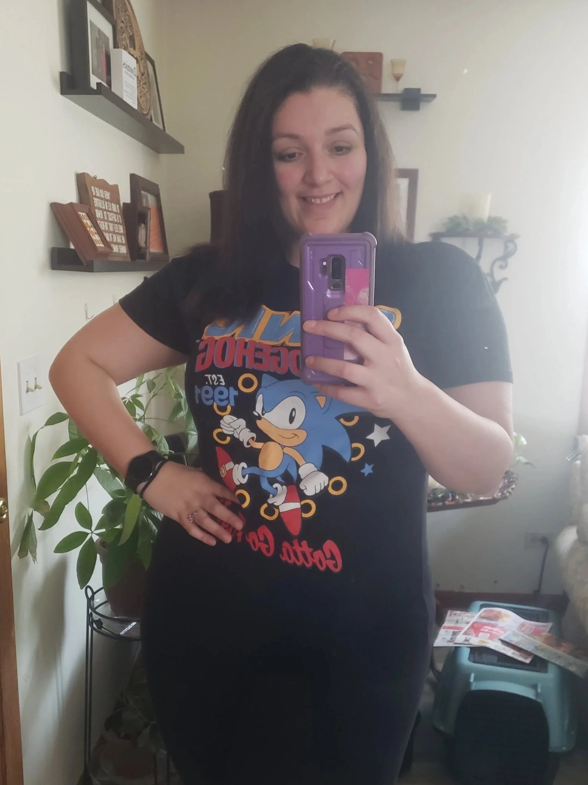 A woman taking a mirror selfie wearing a black Sonic the Hedgehog graphic t-shirt. She has long dark hair, a smartwatch on her left wrist, and is smiling. The room has a plant, shelves with decorations, and a small table with magazines or flyers.