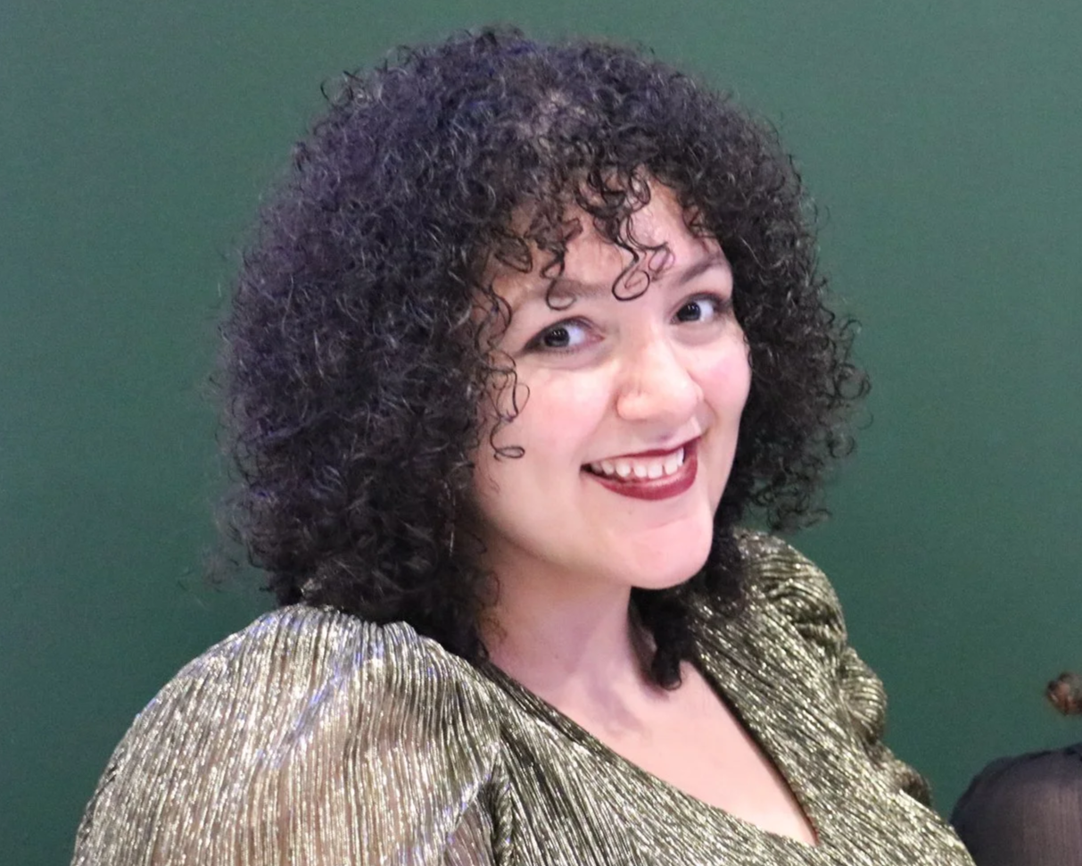 A woman with curly dark hair, wearing a shiny gold top, smiling against a green background.