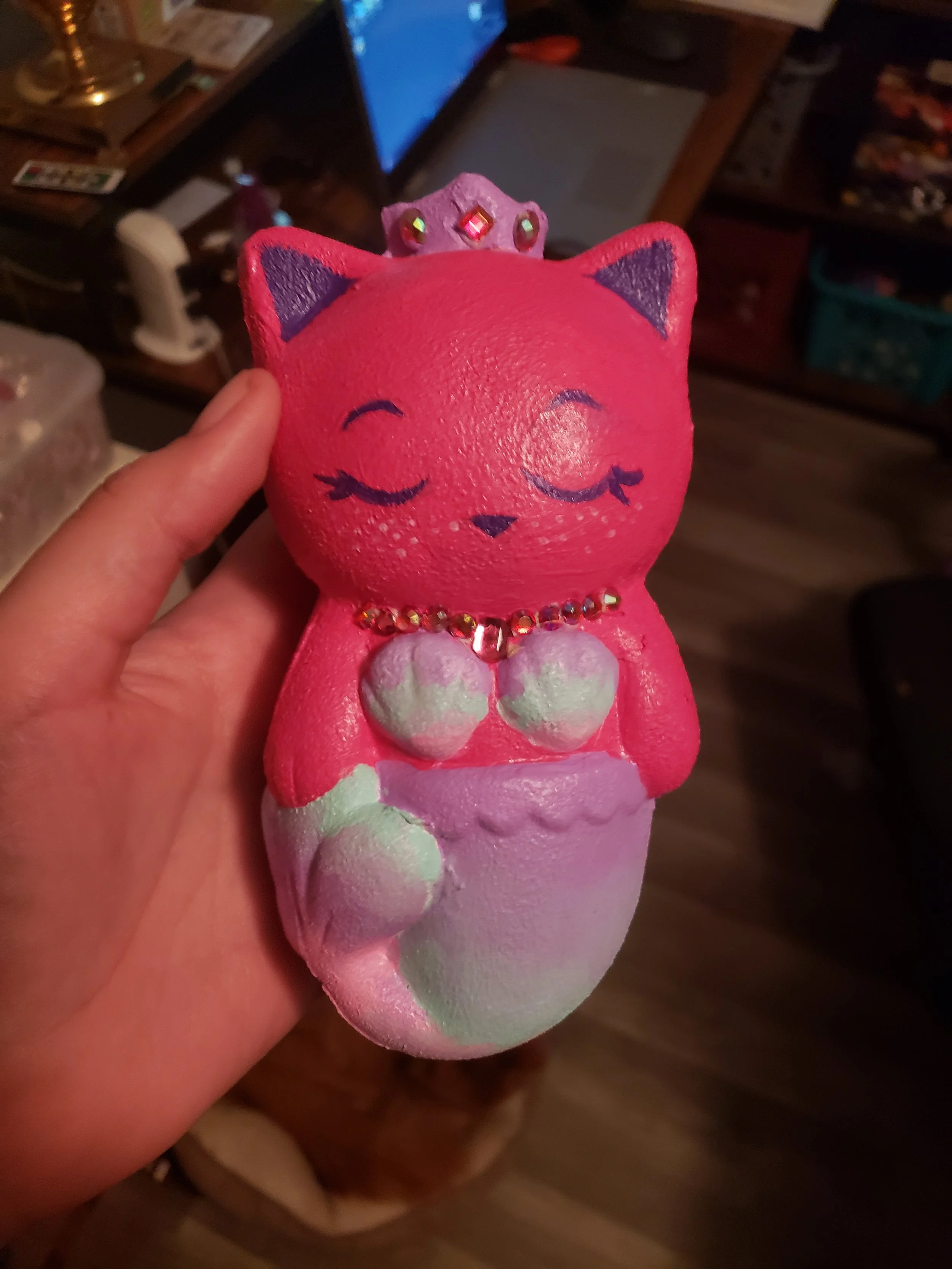 Pink and purple mermaid-shaped figurine of a cat with a crown on its head, closed eyes, and a necklace with rhinestones.