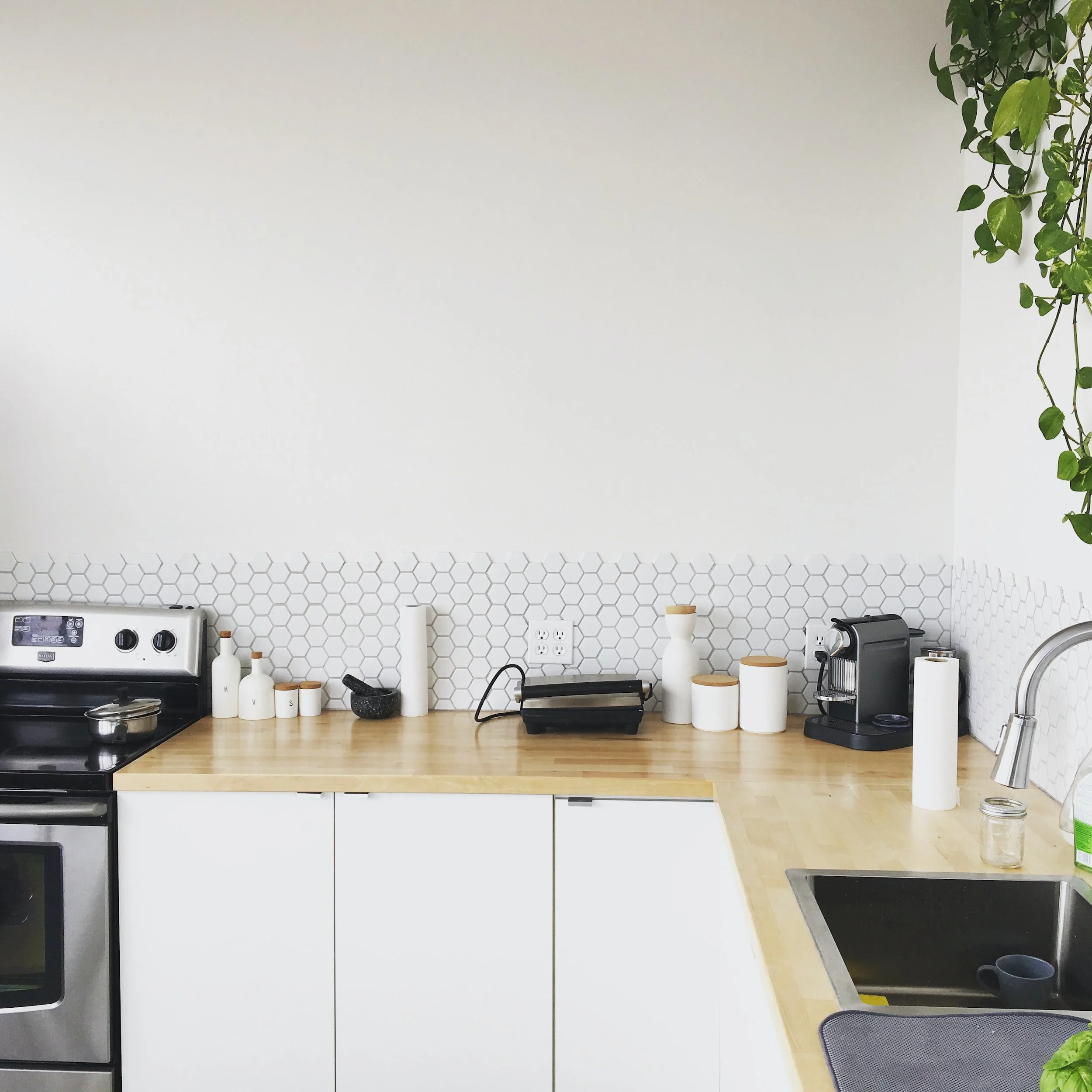 Kitchen with white cabinets, a wooden countertop, a black stove, a coffee machine, and decorative ceramic jars, with a white hexagon-tile backsplash and a small plant hanging from the ceiling.