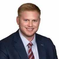 Professional headshot of a man with short blond hair, dressed in a navy suit, white shirt, and striped red and blue tie, smiling against a plain white background.