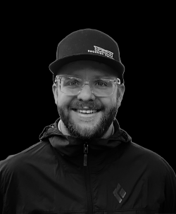 A man with glasses, a beard, and a smiling face wearing a black cap and a black jacket, set against a plain background.
