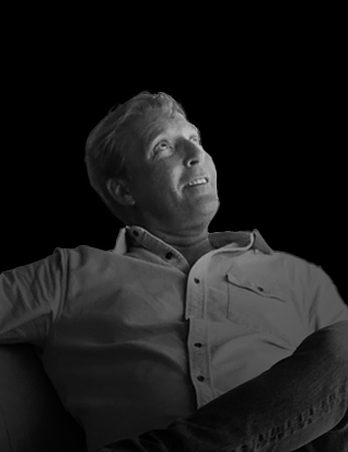 A black and white photo of a man looking upward, sitting with legs crossed, wearing a collared shirt.