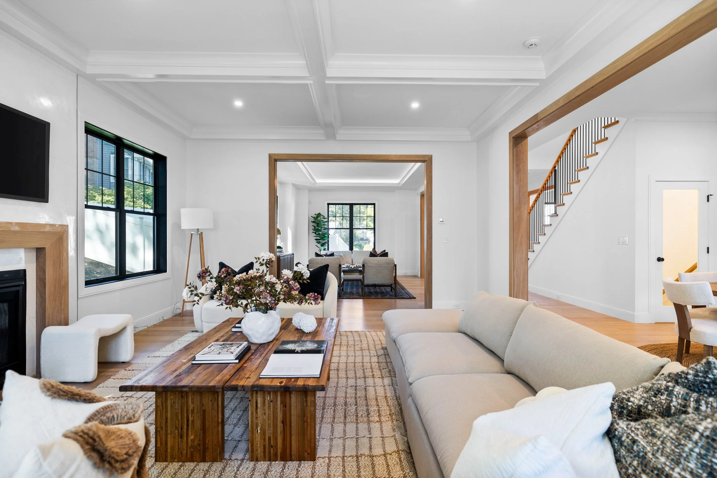 A modern living room with white walls, hardwood floors, and a large beige sofa. There is a wooden coffee table with books and a vase of flowers, a white upholstered stool, and a black-framed window. In the background, another room with additional sea