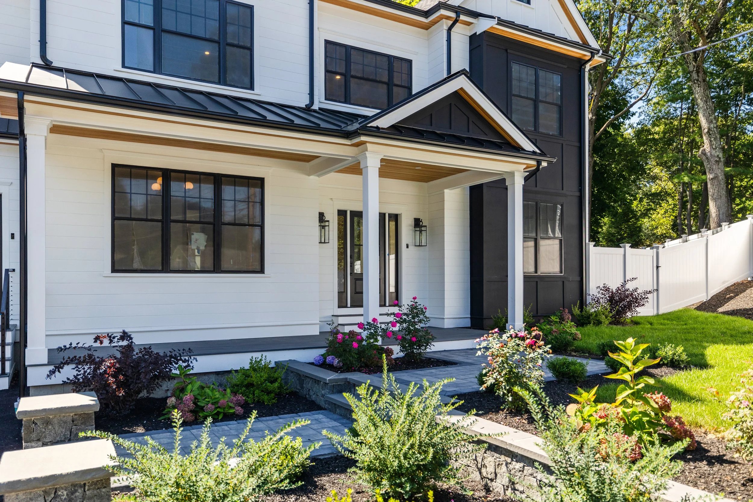 Modern house with white exterior, black window frames, and a black roof. Small porch with white columns, landscaped garden with colorful flowers and shrubs, white fence, and surrounding green trees.