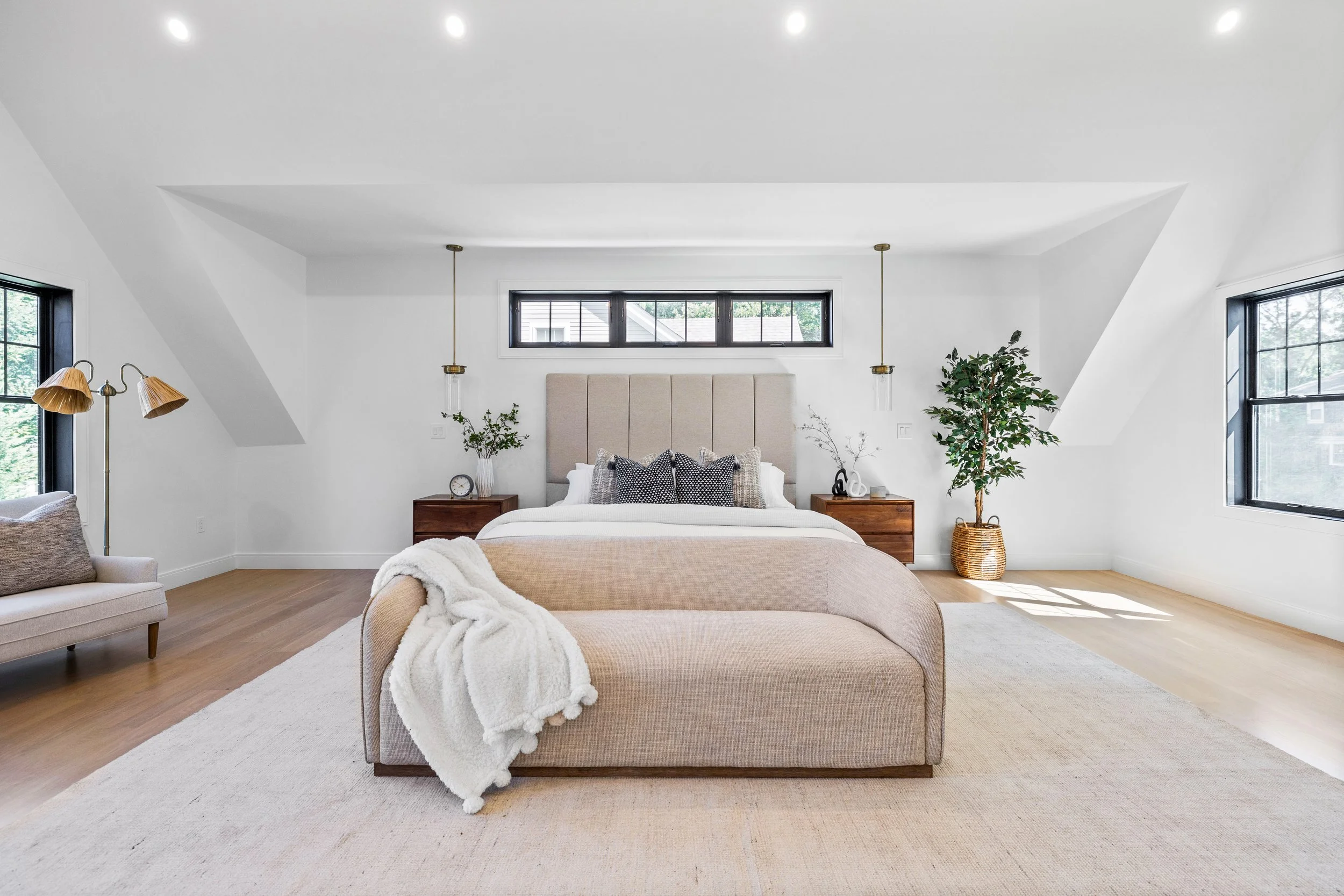Bright and minimalist bedroom interior with a central bed, beige headboard, and extra seating. The room has large black-framed windows, light wood flooring, a white rug, and decorative plants.