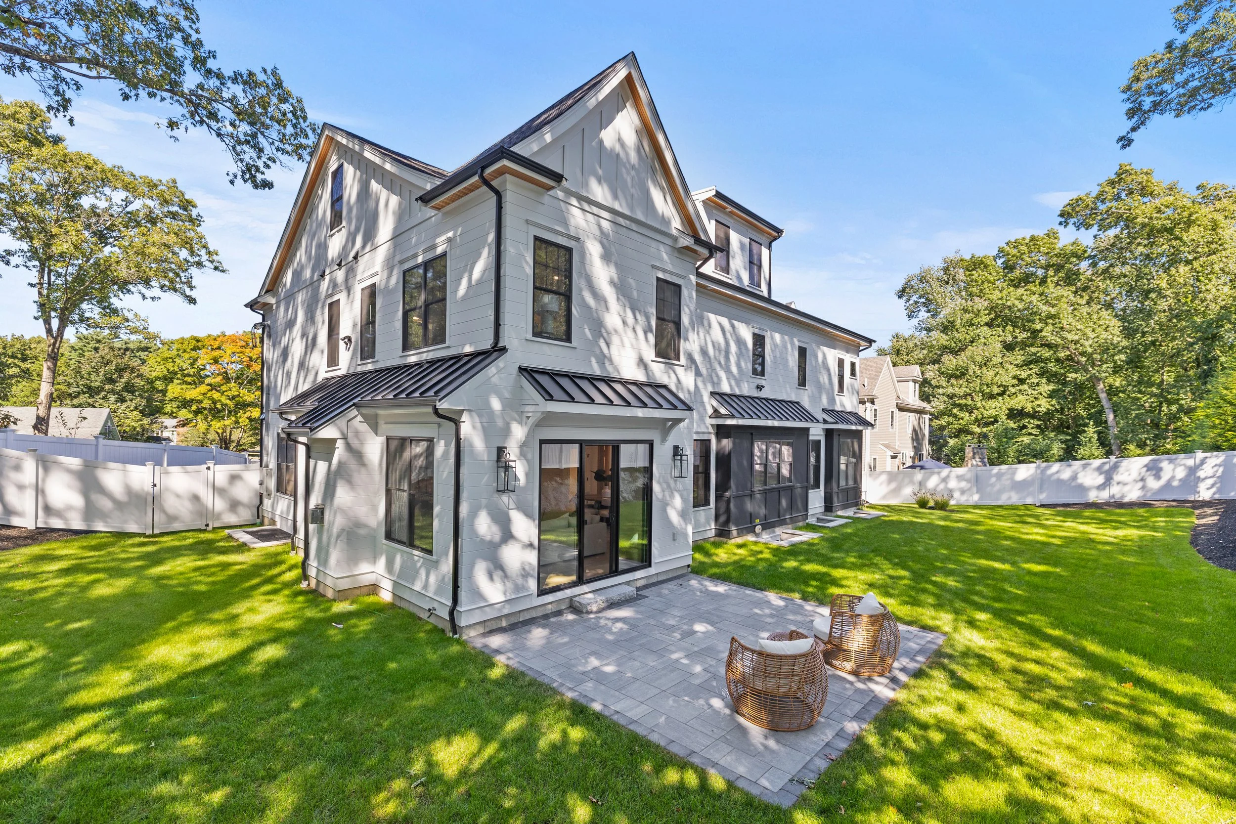 White modern house with a backyard, patio, and green lawn, surrounded by a white fence and mature trees under a blue sky.