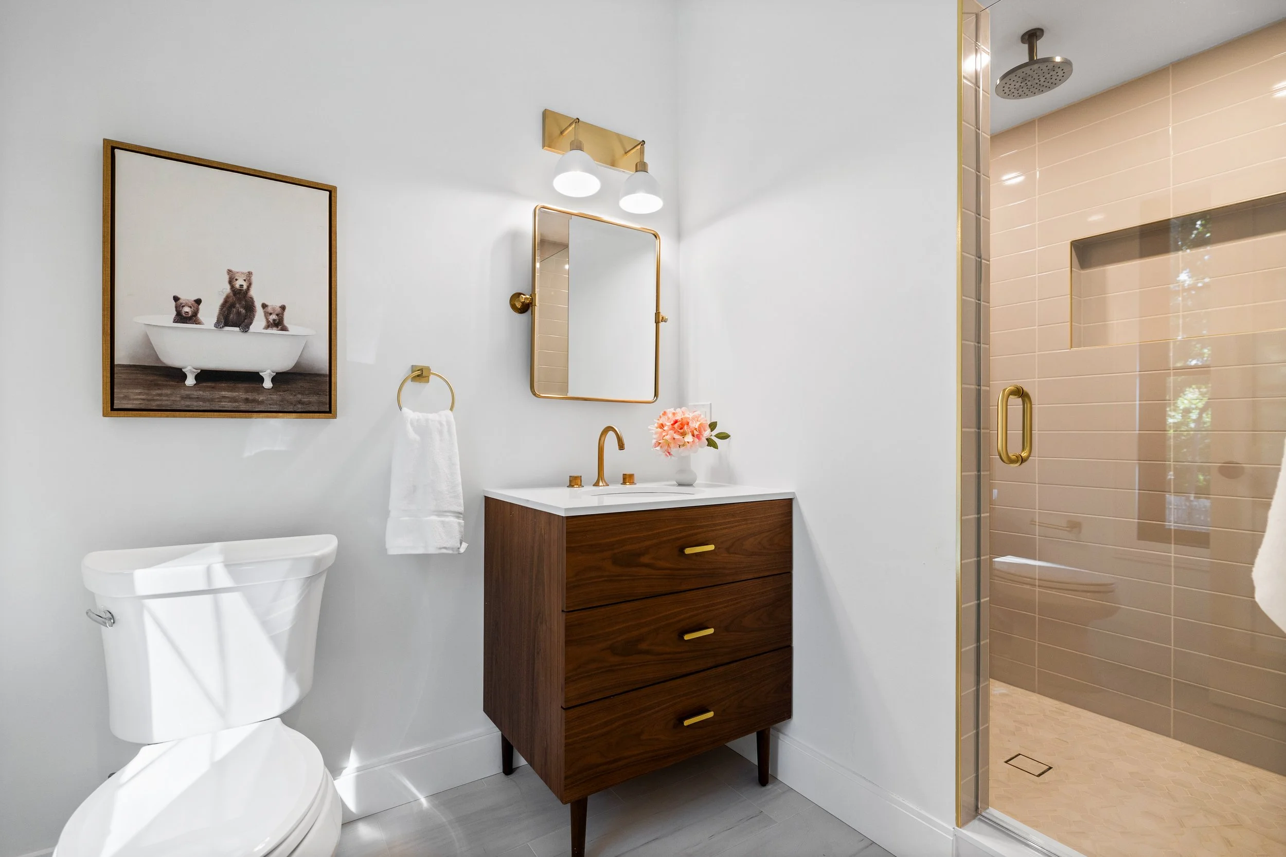 Modern bathroom with white walls, a wooden vanity with gold hardware and a sink, a gold-framed mirror, a wall-mounted light fixture, a framed picture of bears in a bathtub, a white towel on a gold ring, a white toilet, and a walk-in shower with beige